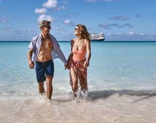 Couple walking on a beach in the Caribbean, with a Holland America cruise ship on the horizon.