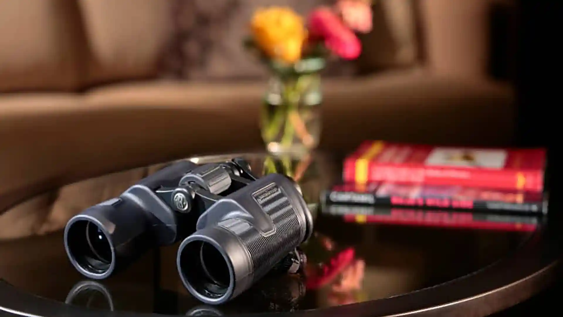 Binoculars on brown wooden table with books and flowers in background.