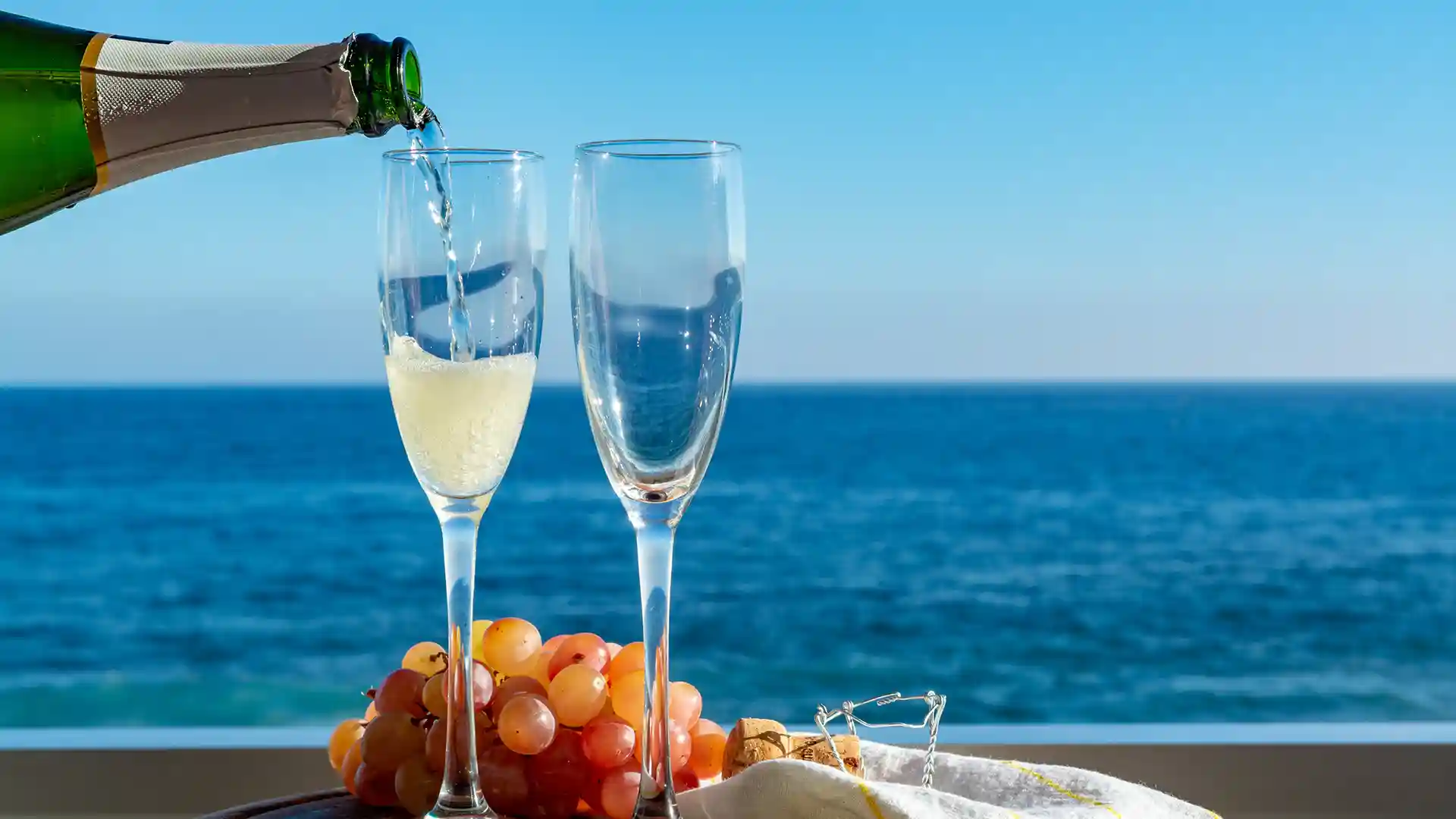 View of champagne being poured into glasses next to grapes on table with ocean in background.