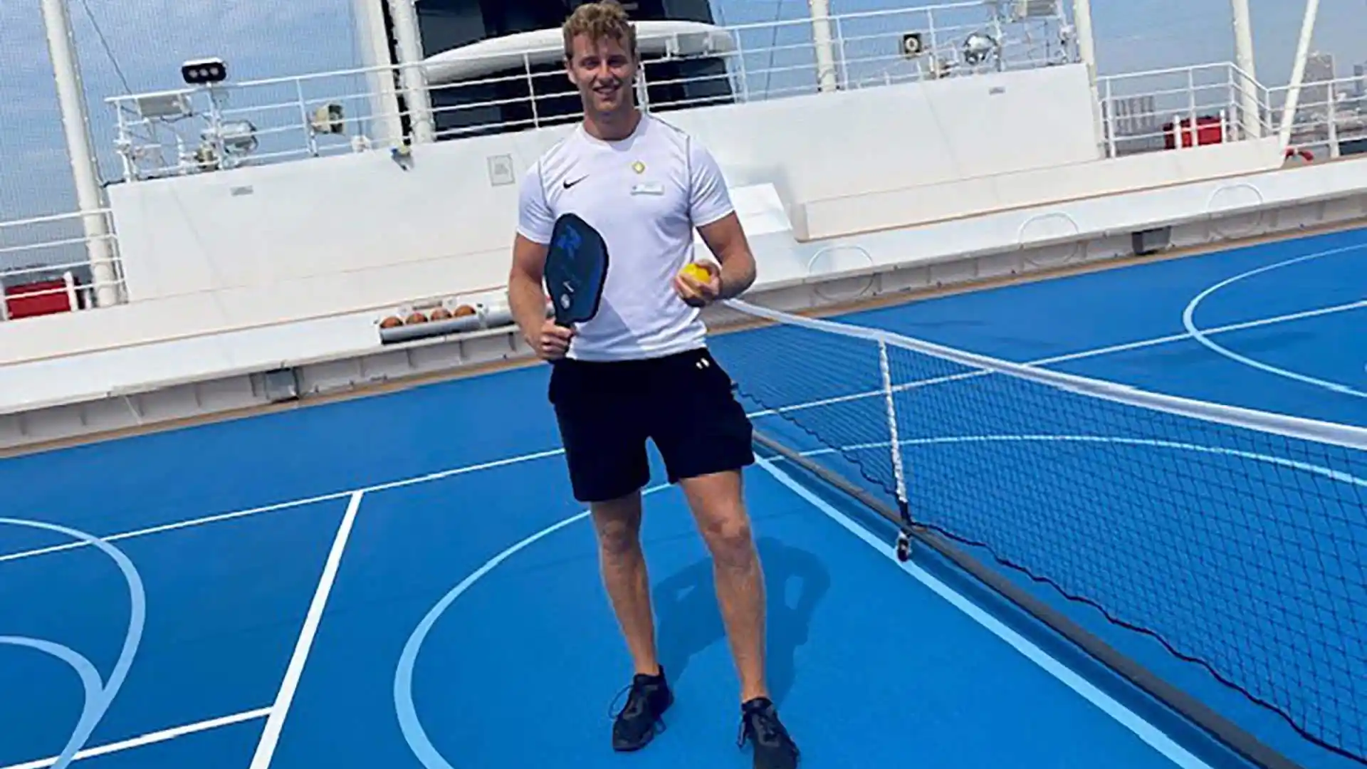 Person holding pickleball paddle and serving ball on court on Holland America Line cruise ship.