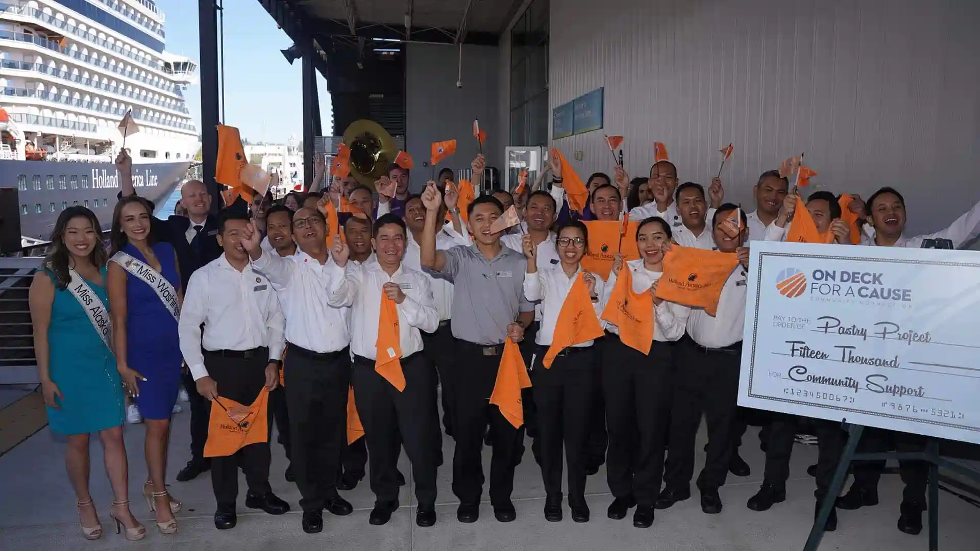 View of people holding orange Holland America Line flags, waving in celebration at cruise port with cruise ship in background.