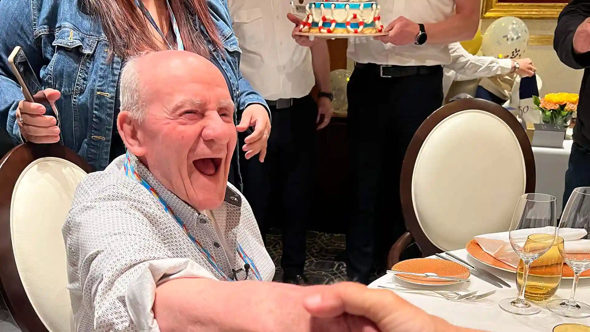 Person smiling and shaking hands from dining table with people standing in background.