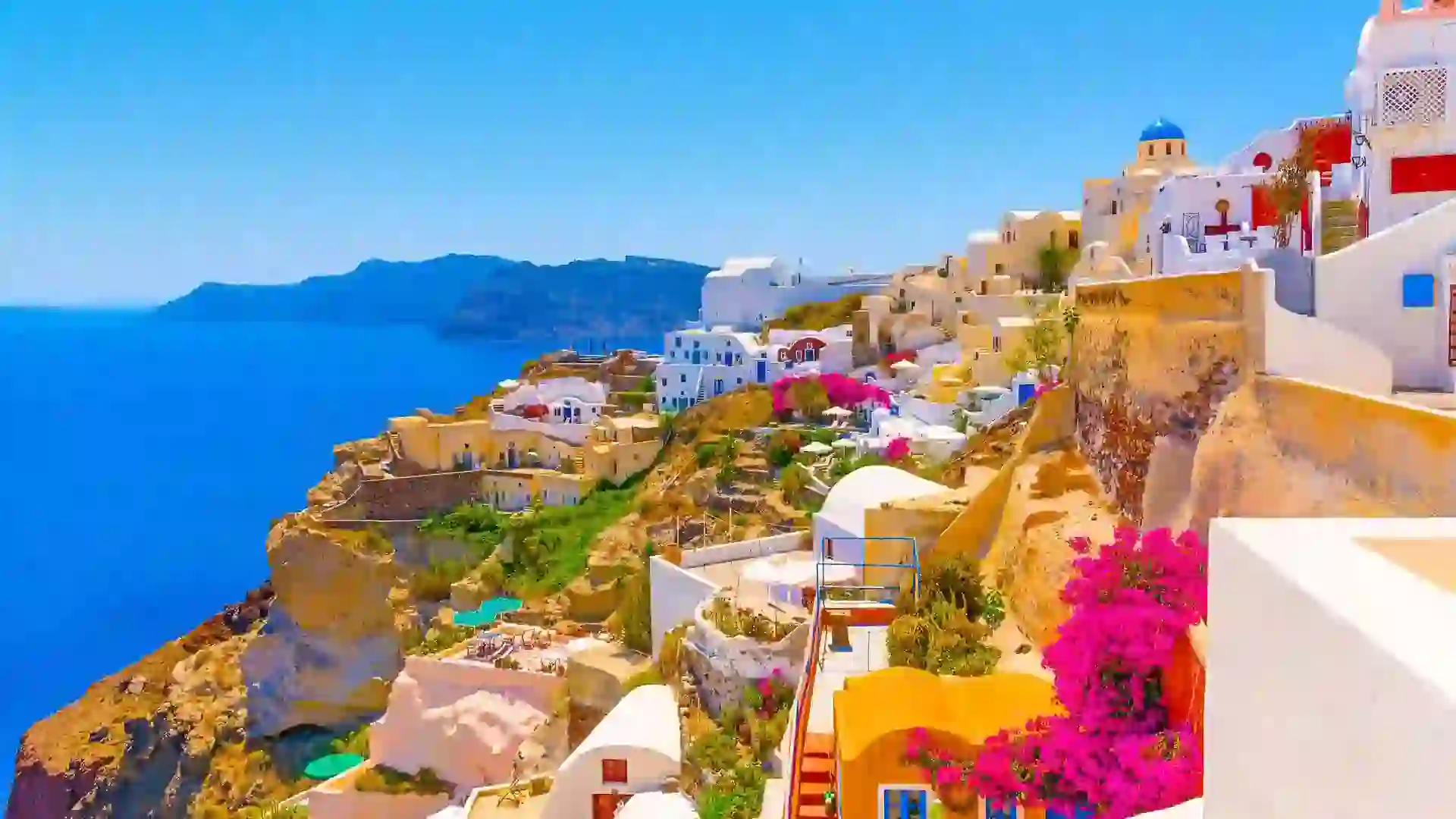 White and orange buildings cover hillside with green landscape and blue ocean water in background.