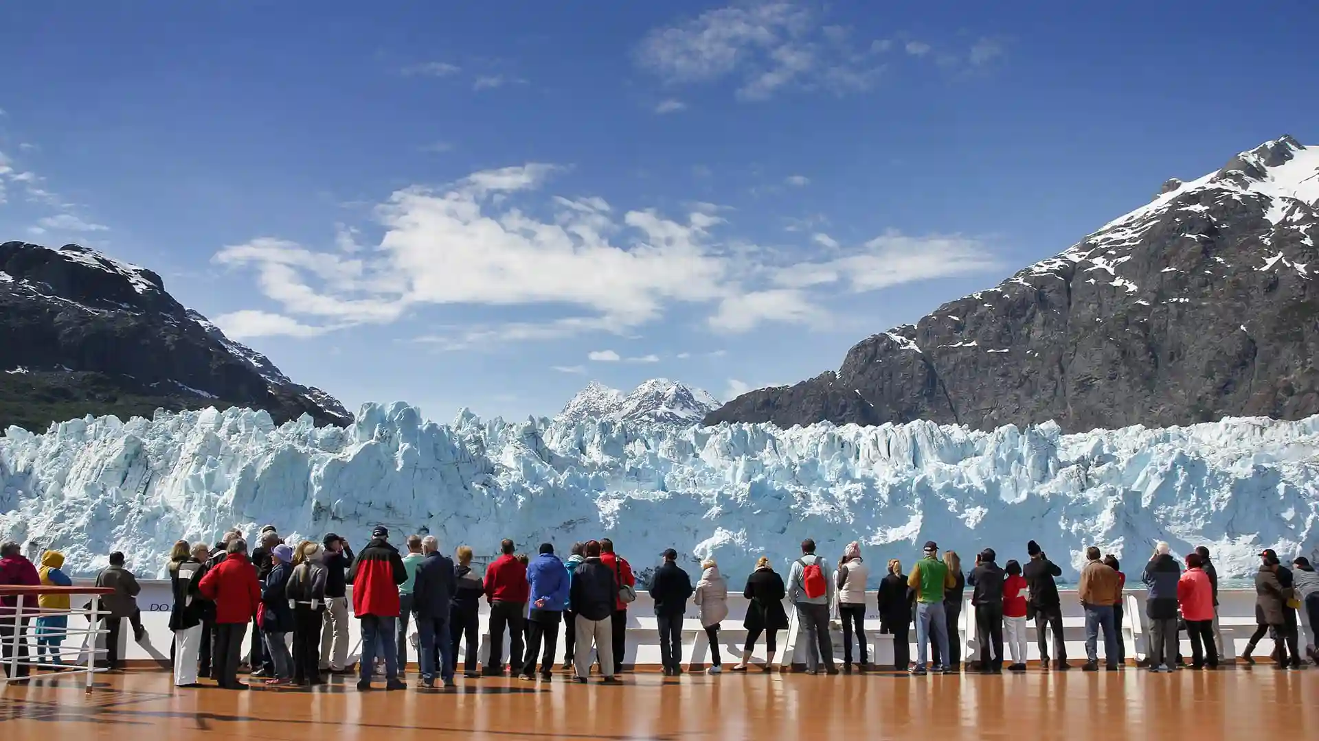People on cruise deck viewing icy glacier beneath blue sky.