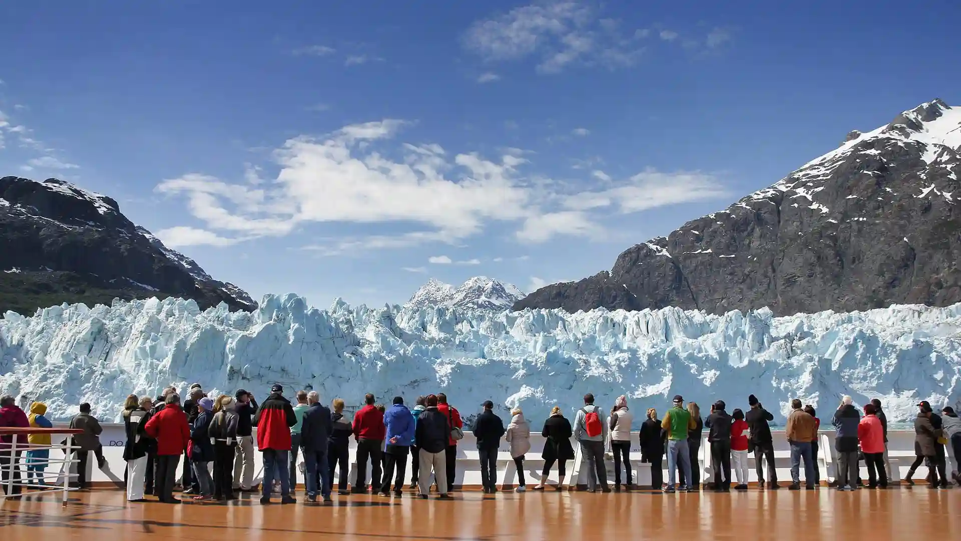 Alaska Glacier Views People on cruise deck viewing icy glacier beneath blue sky.