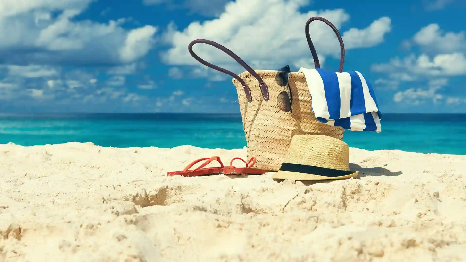 Beach bag with towel, sunglasses, hat and flipflops on beach.
