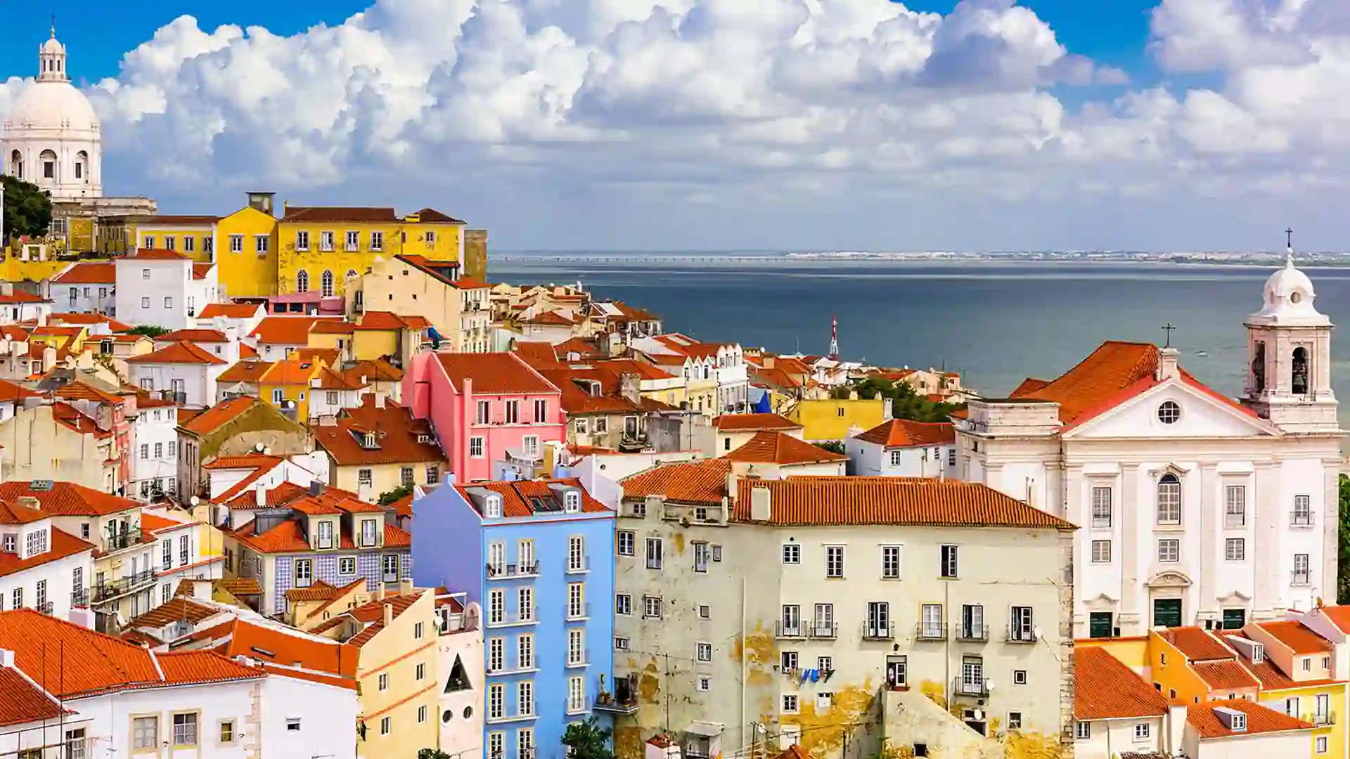 View of white, orange and blue buildings with red roofs and the ocean and cloudy skies in background.