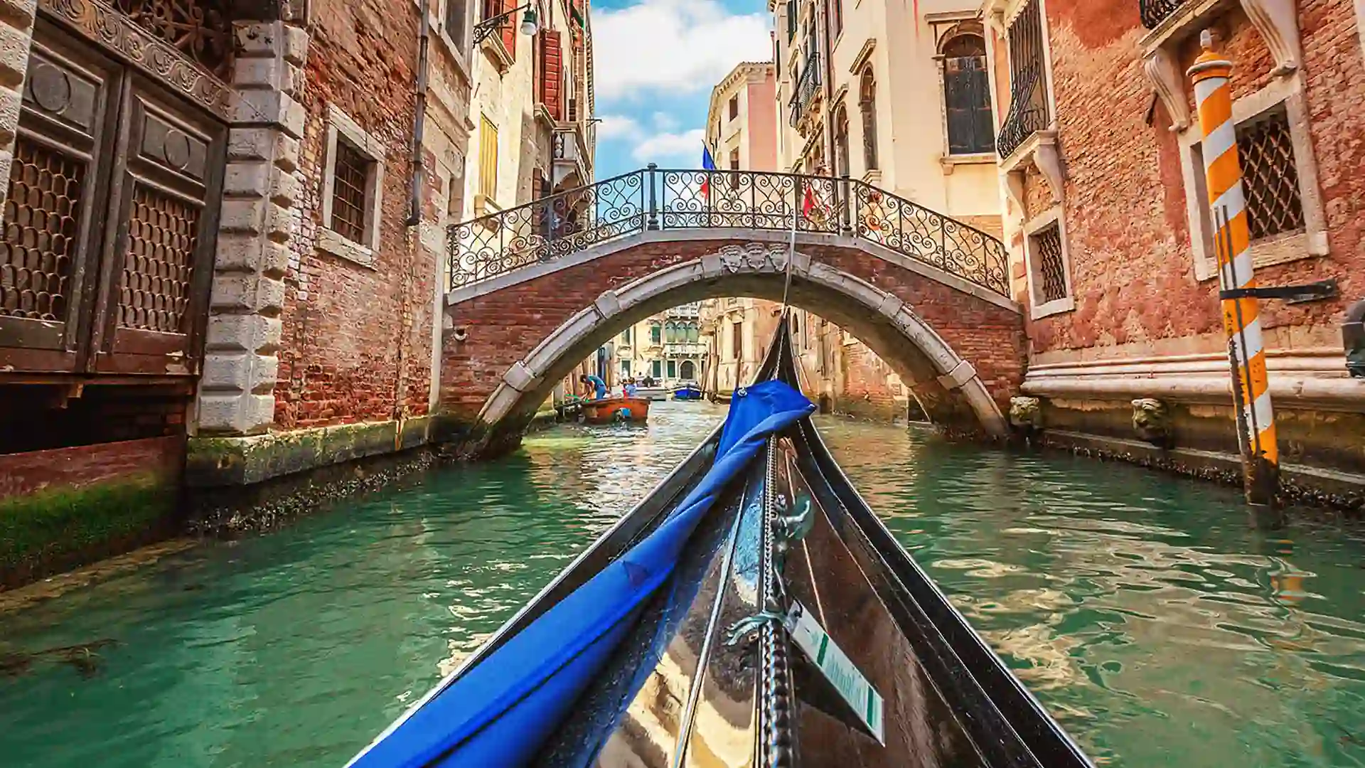 View from bow of gondola sailing toward bridge on narrow waterway surrounded by buildings.