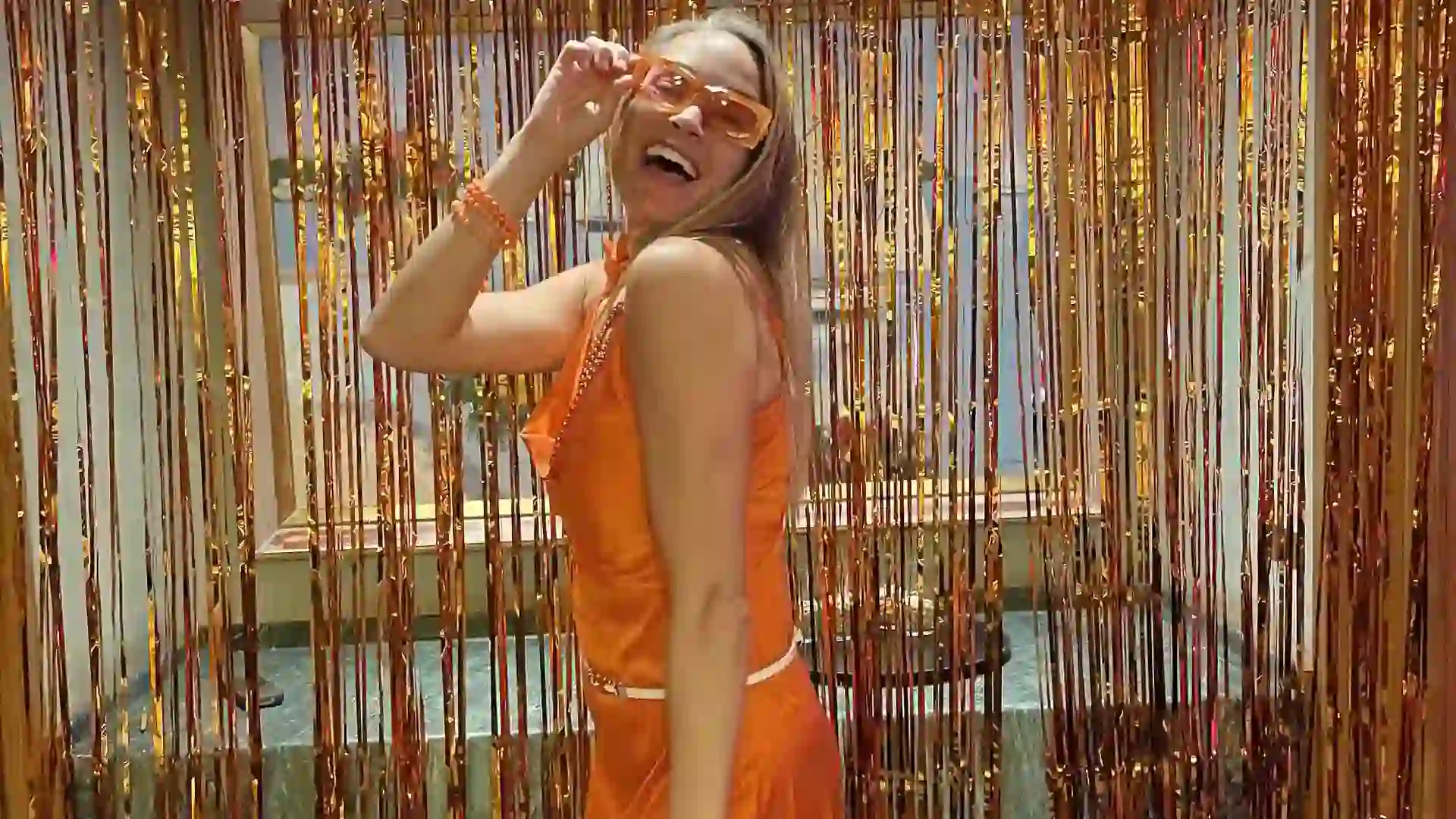 Person wearing orange dress and sunglasses in front of orange streamers at party.