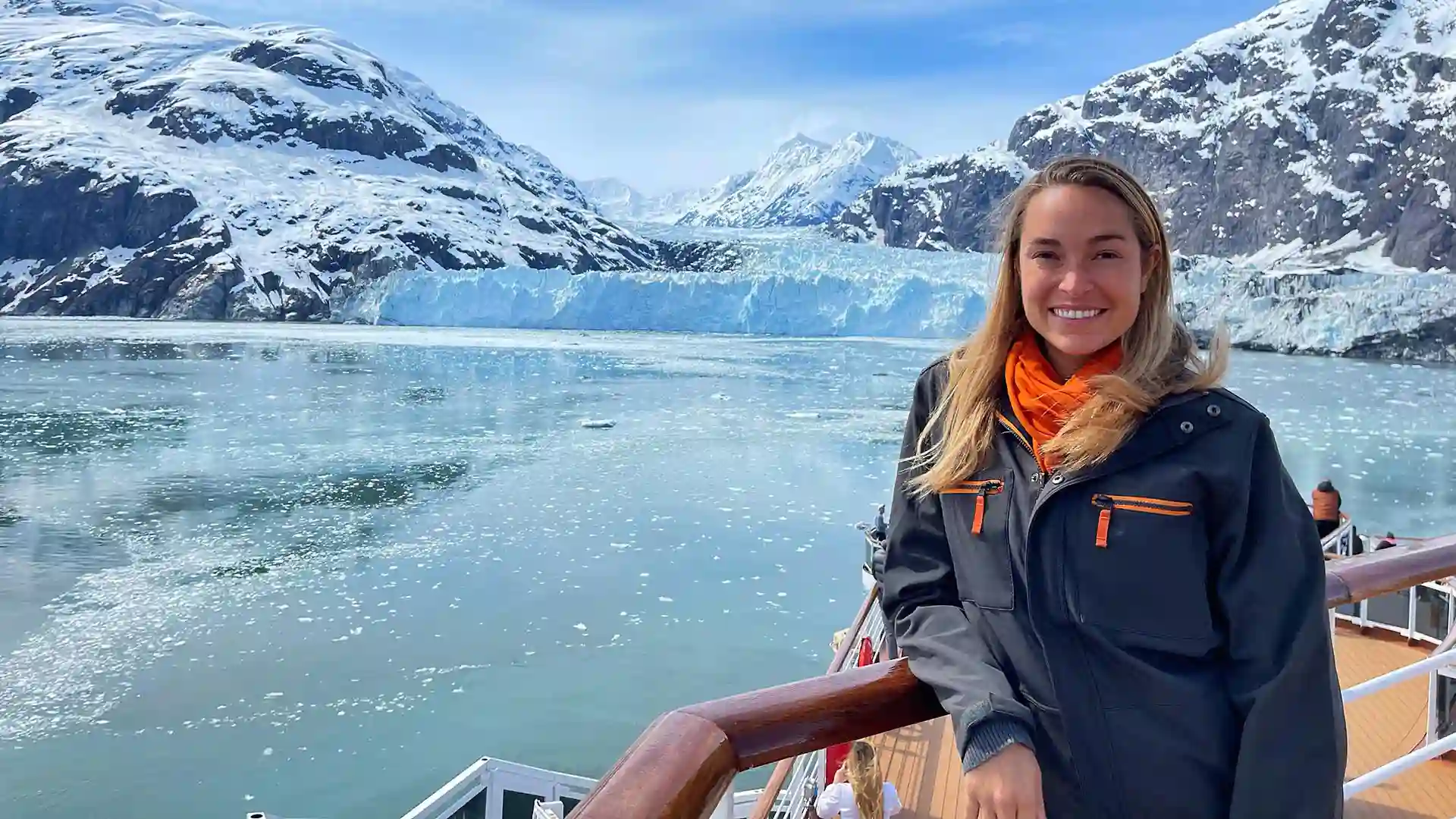 Person wearing blue and orange jacket, standing on cruise deck with glacier in background.