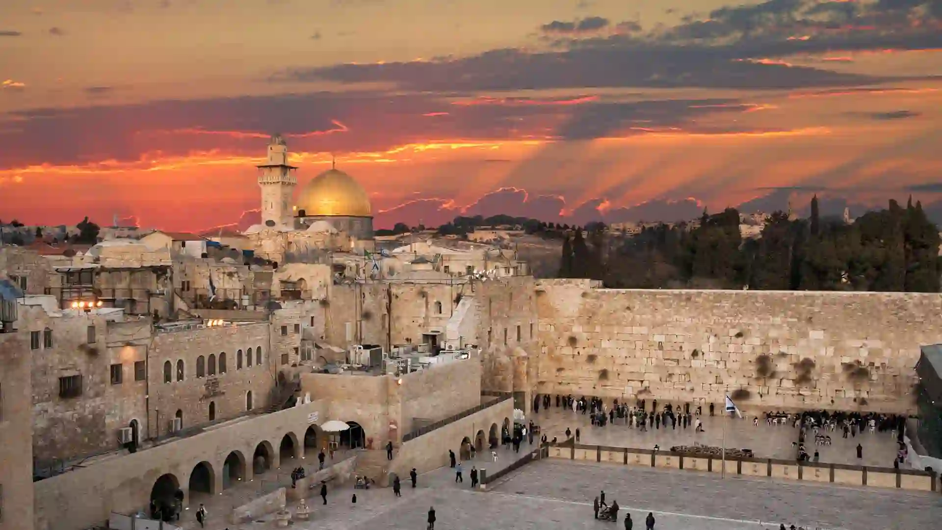 Crowd visits ancient stone landmark with surrounding buildings and temples while sun sets.