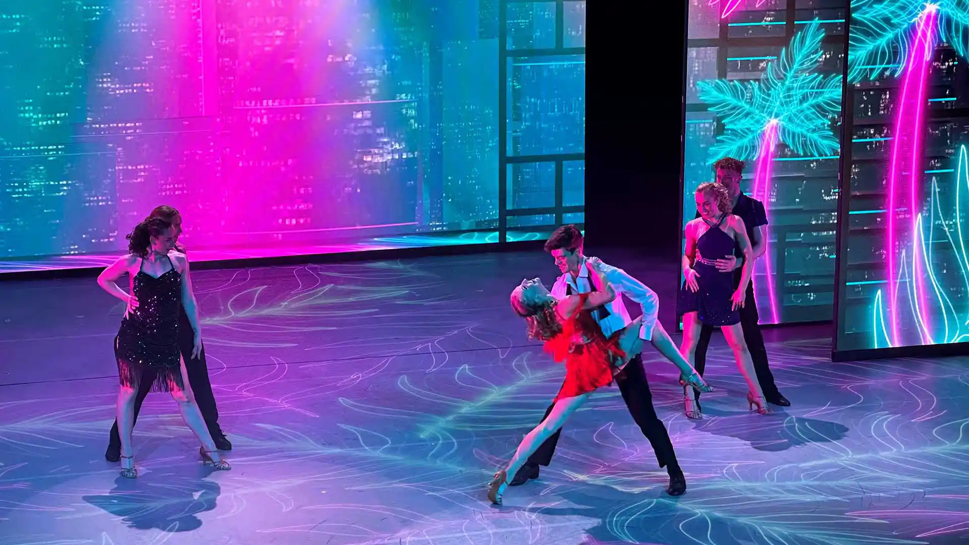 Six dancers perform on stage with teal and pink lighting above and behind them.