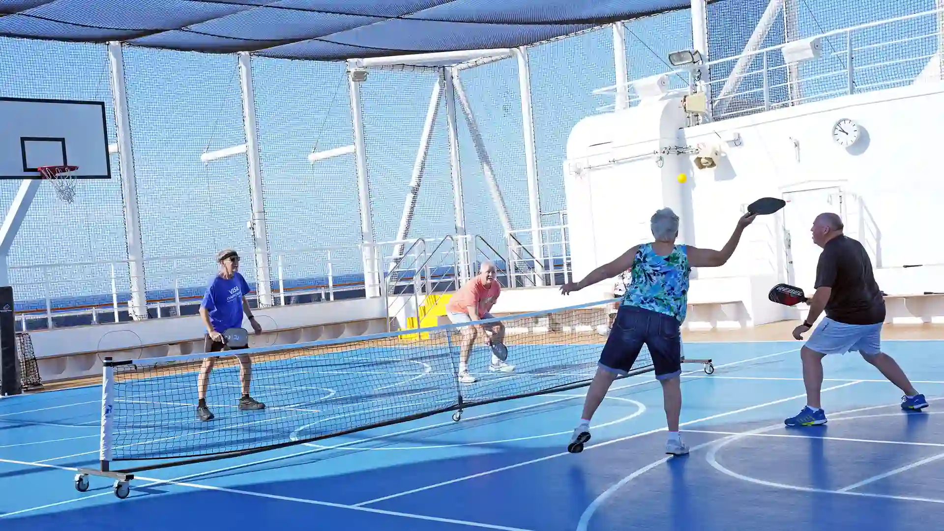 Pickleball Onboard Four people playing pickleball on blue court with ocean in background.