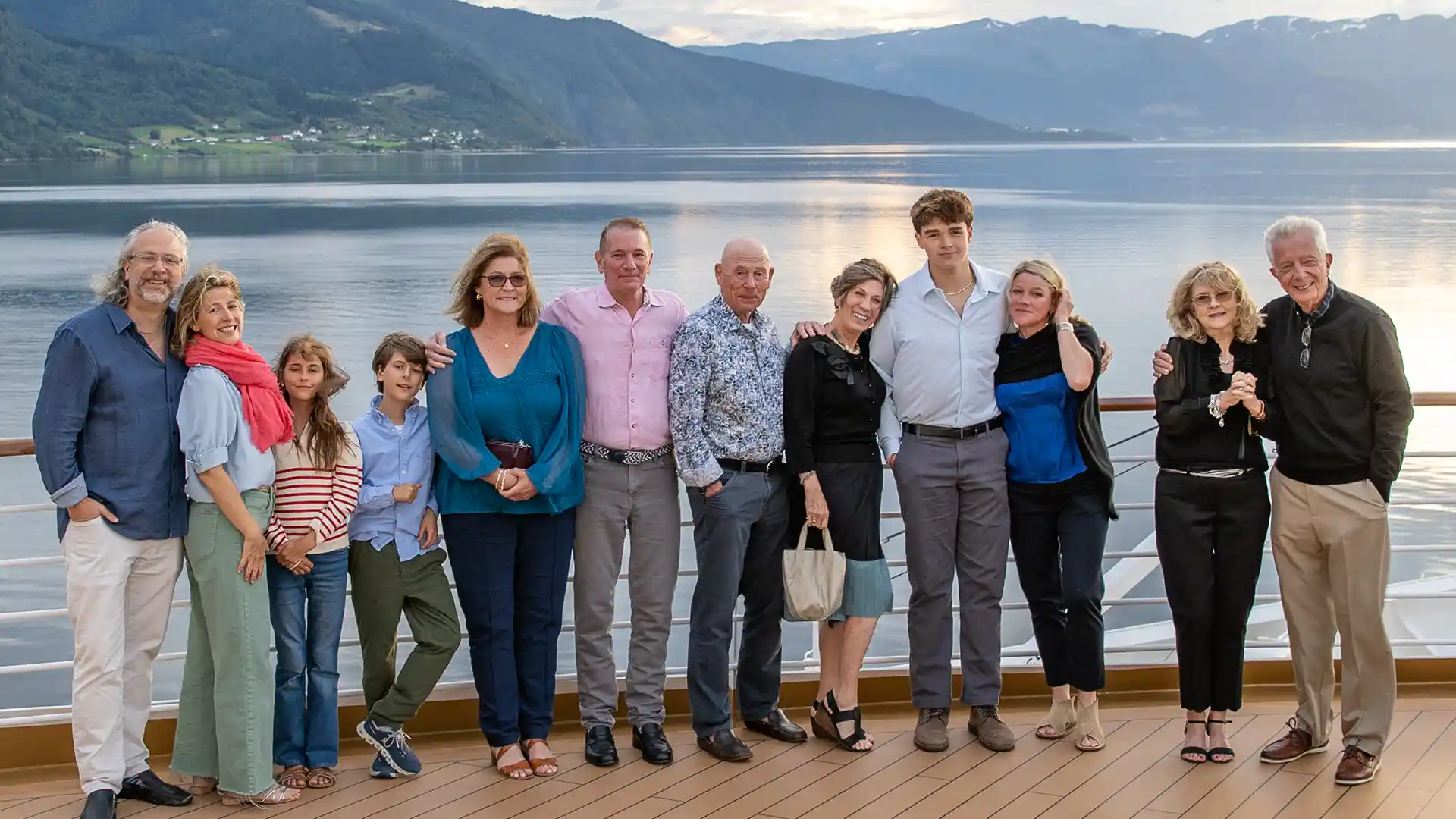 People posing for group photo on cruise ship deck with ocean and green landscape in the background.