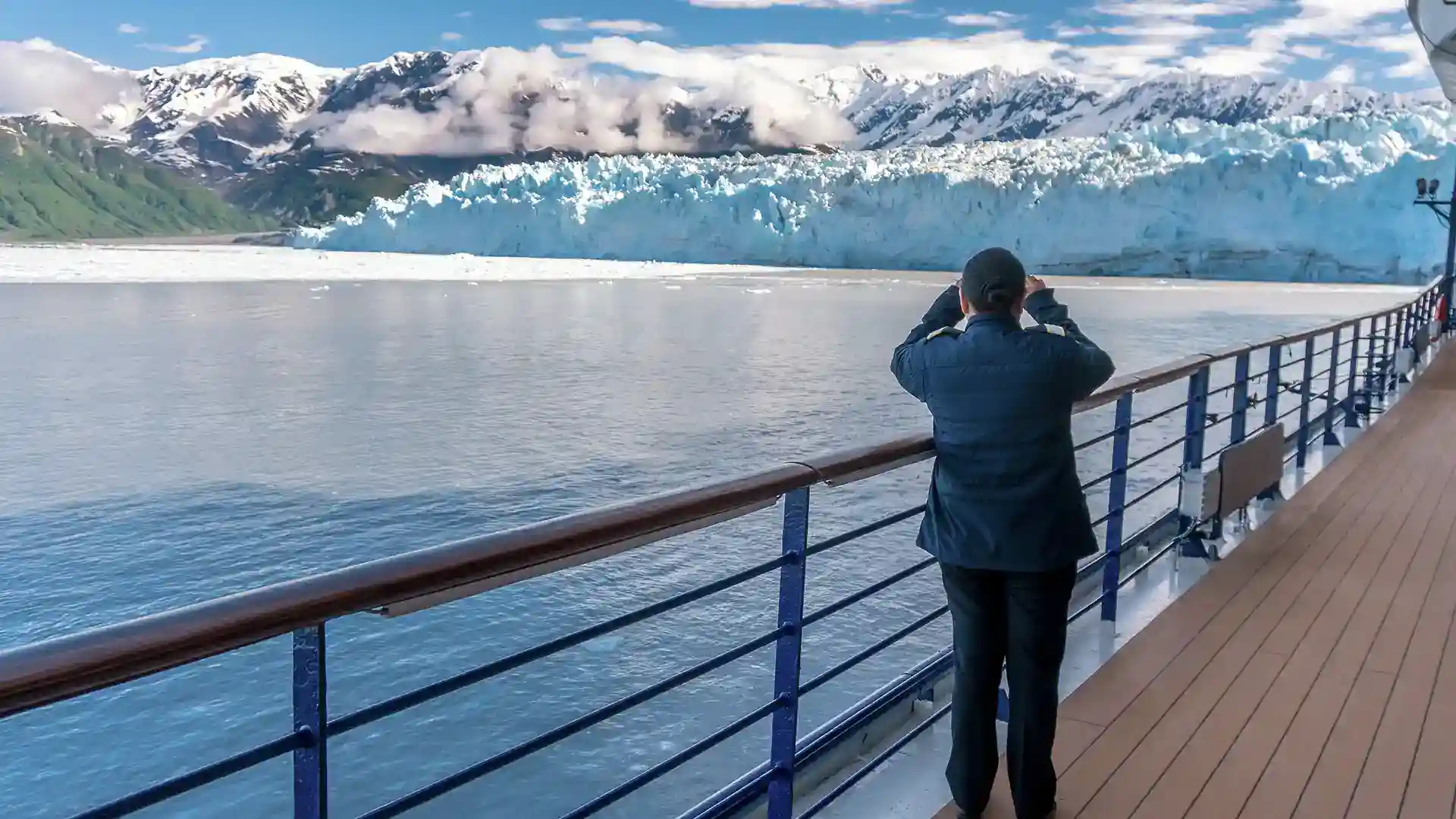 Person in blue jacket on ship deck looking at glacier through binoculars.