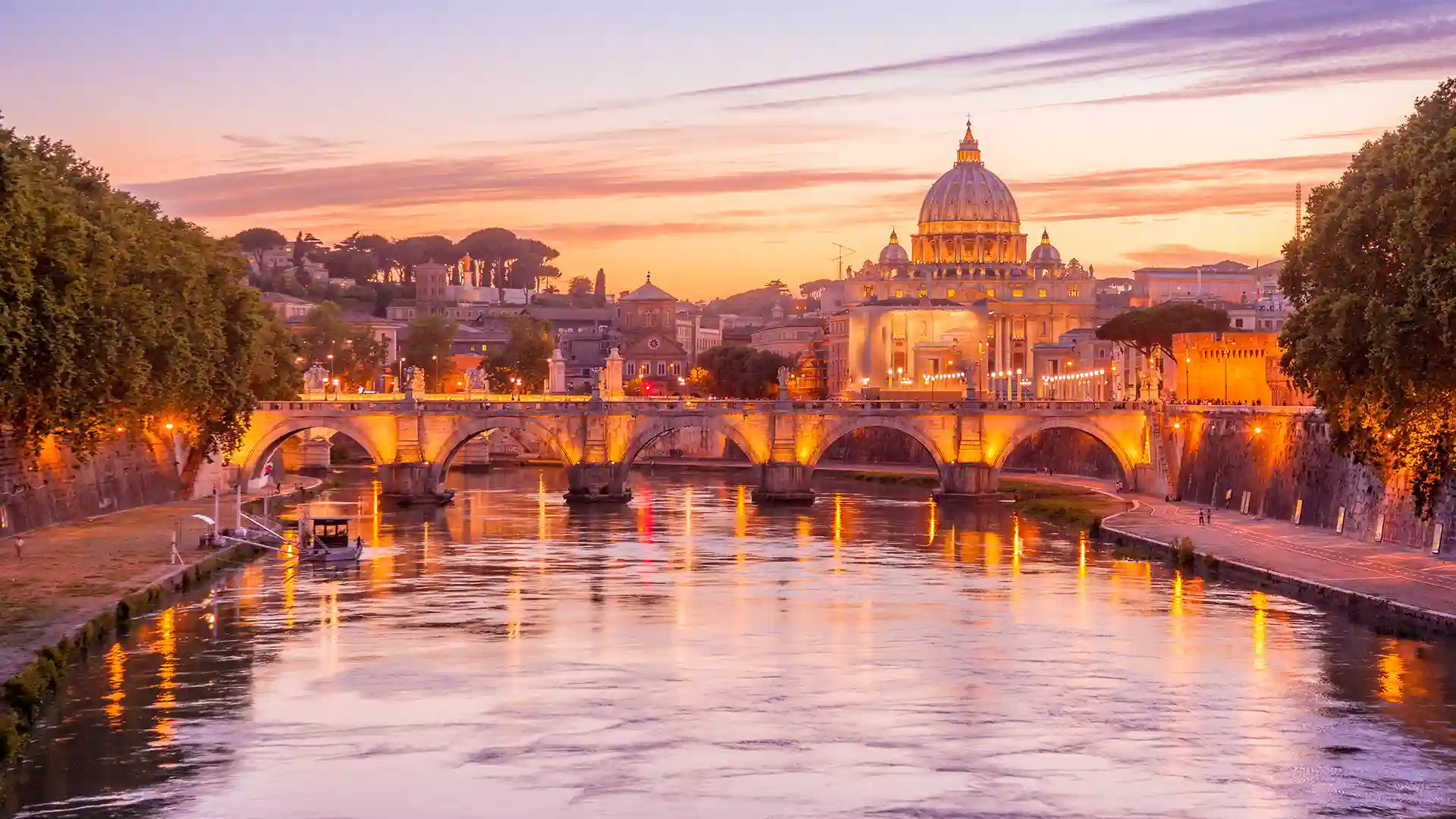 View of water flowing under bridge and evening lights illuminating tall ancient buildings as the sun sets.