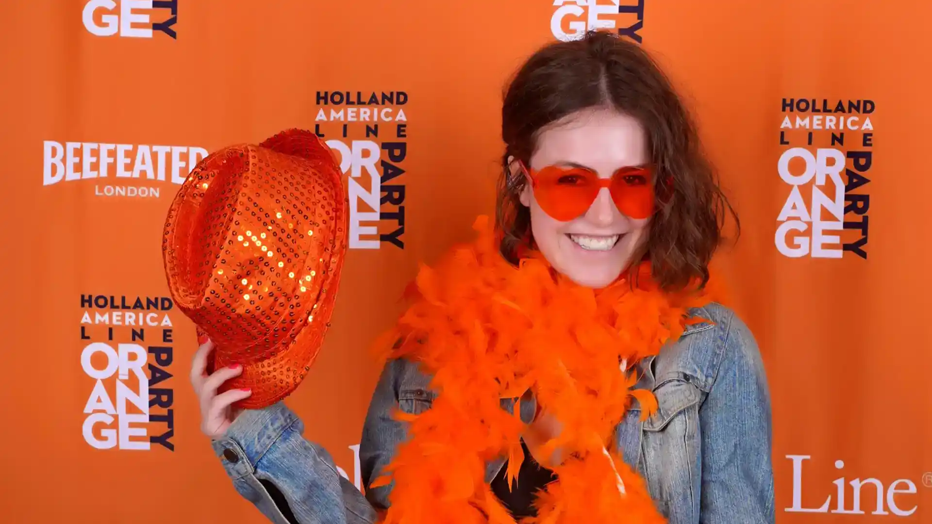 Person wearing orange boa and sunglasses and holding orange hat.