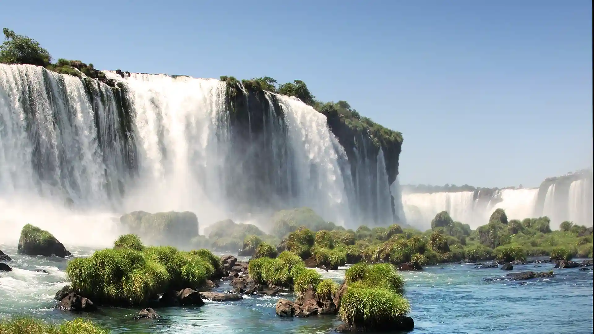 View of Iguazu Falls, a waterfall in South America, surrounded by green landscape and blue sky.
