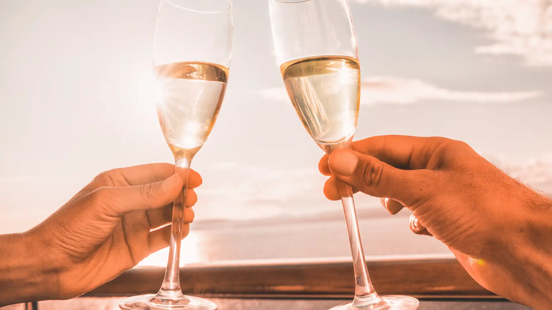 View of hands holding champagne glasses with ocean views and sunset in background.