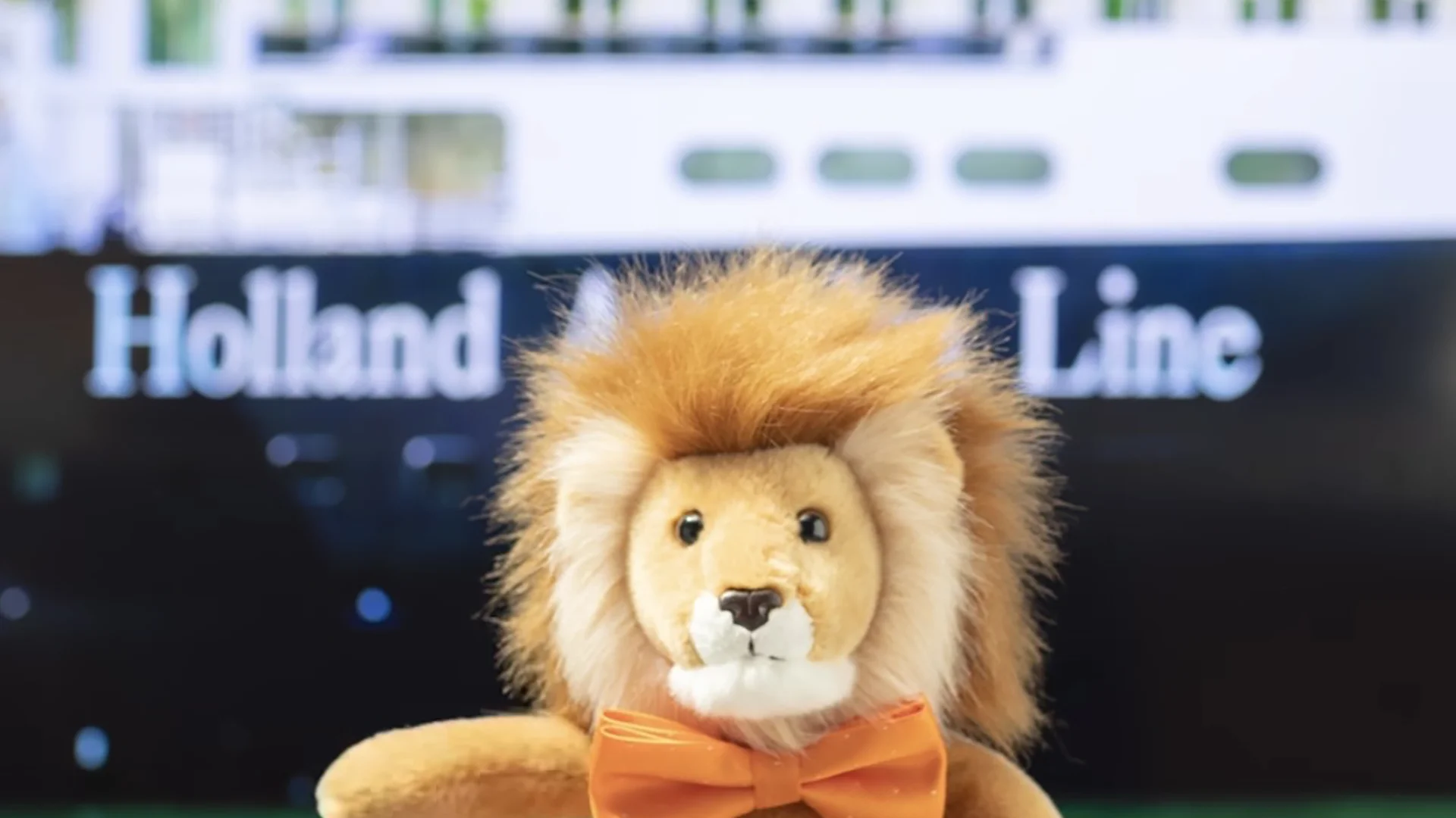 Holland America Line's lion mascot with orange bowtie in front of cruise ship.