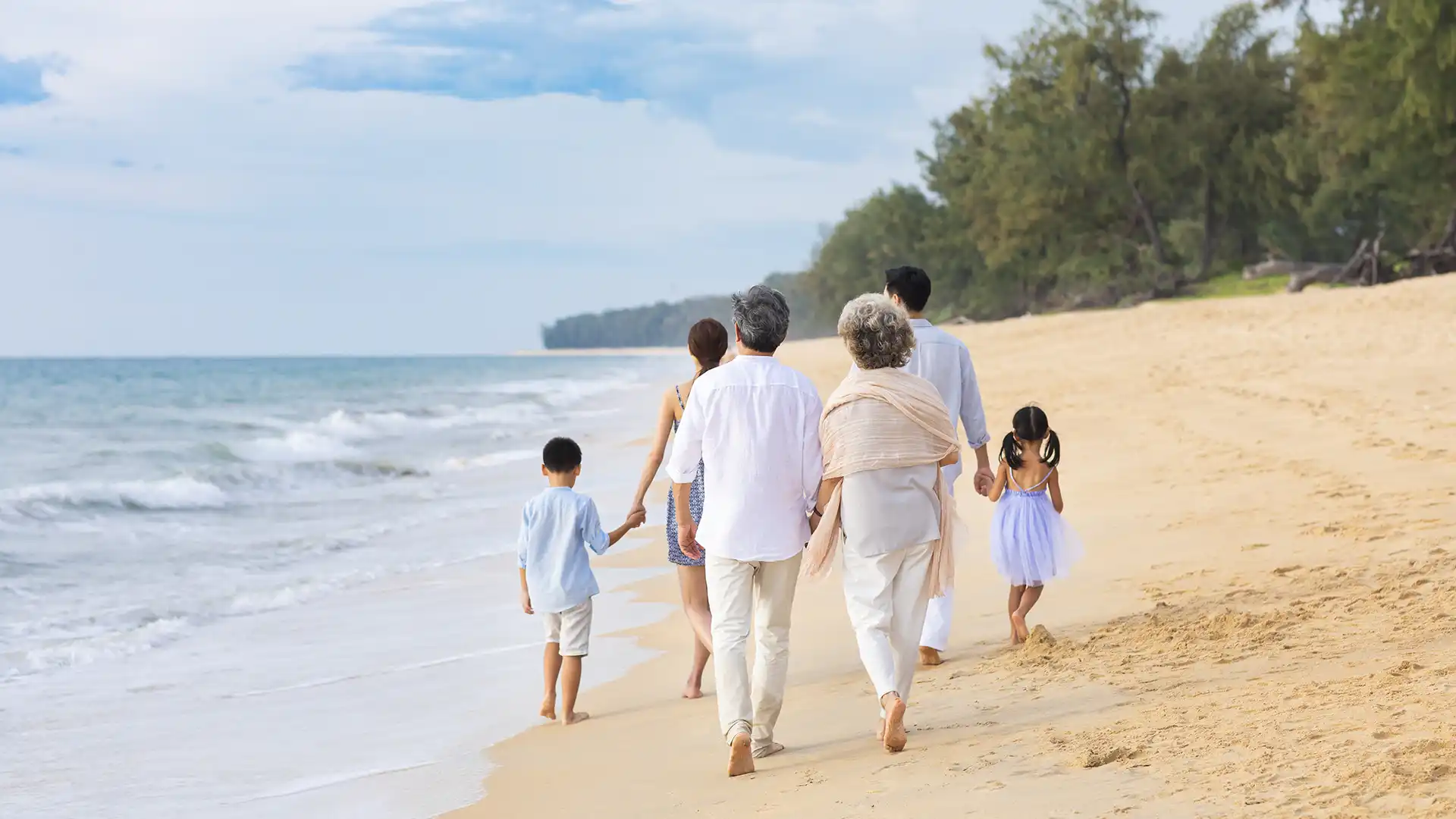 View of adults and children in light-colored clothing walking the beach toward green trees.