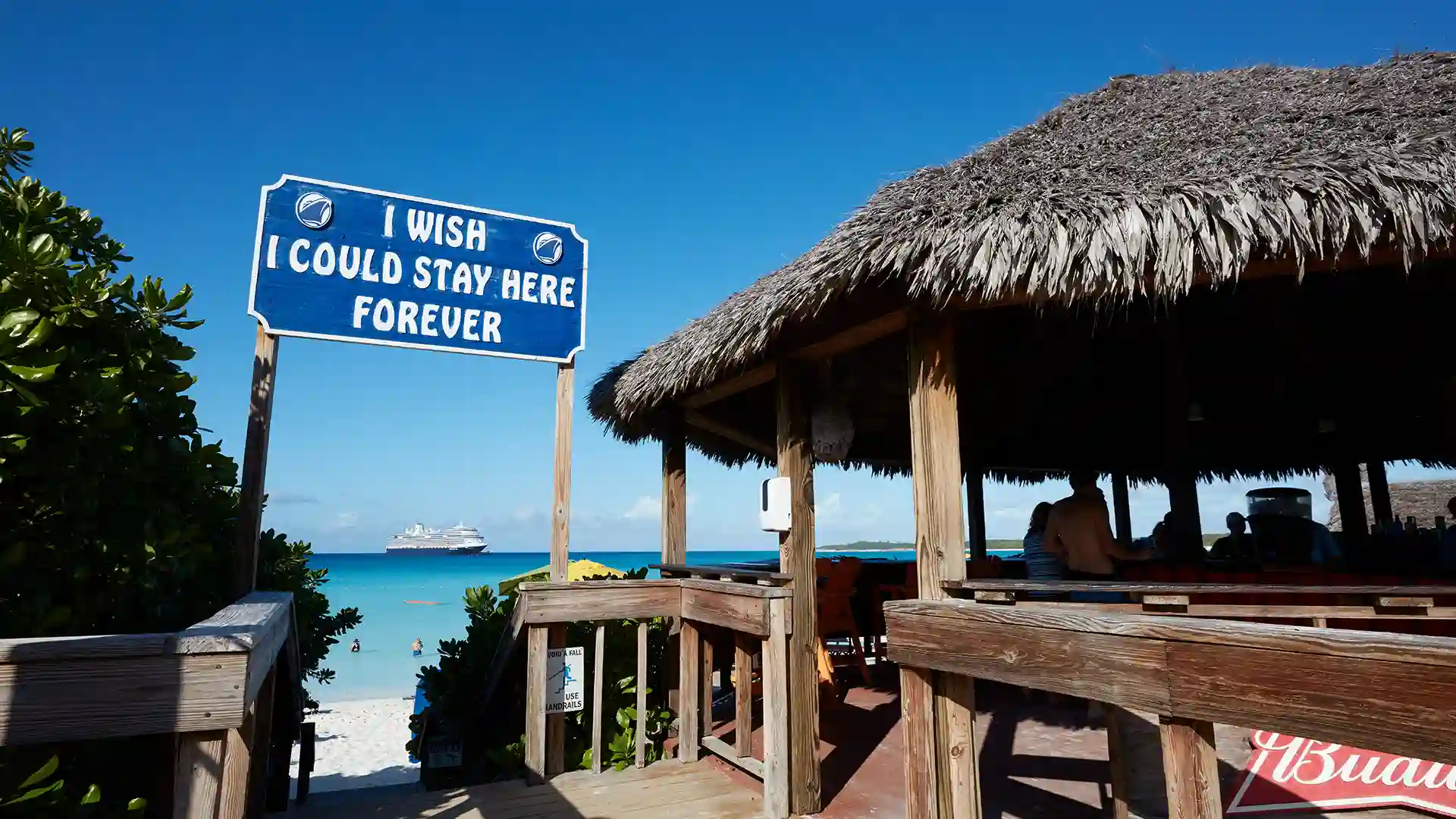 View of cabana and blue and white sign that states "I wish I could stay here forever." Holland America cruise ship appears on bright blue ocean in the background.