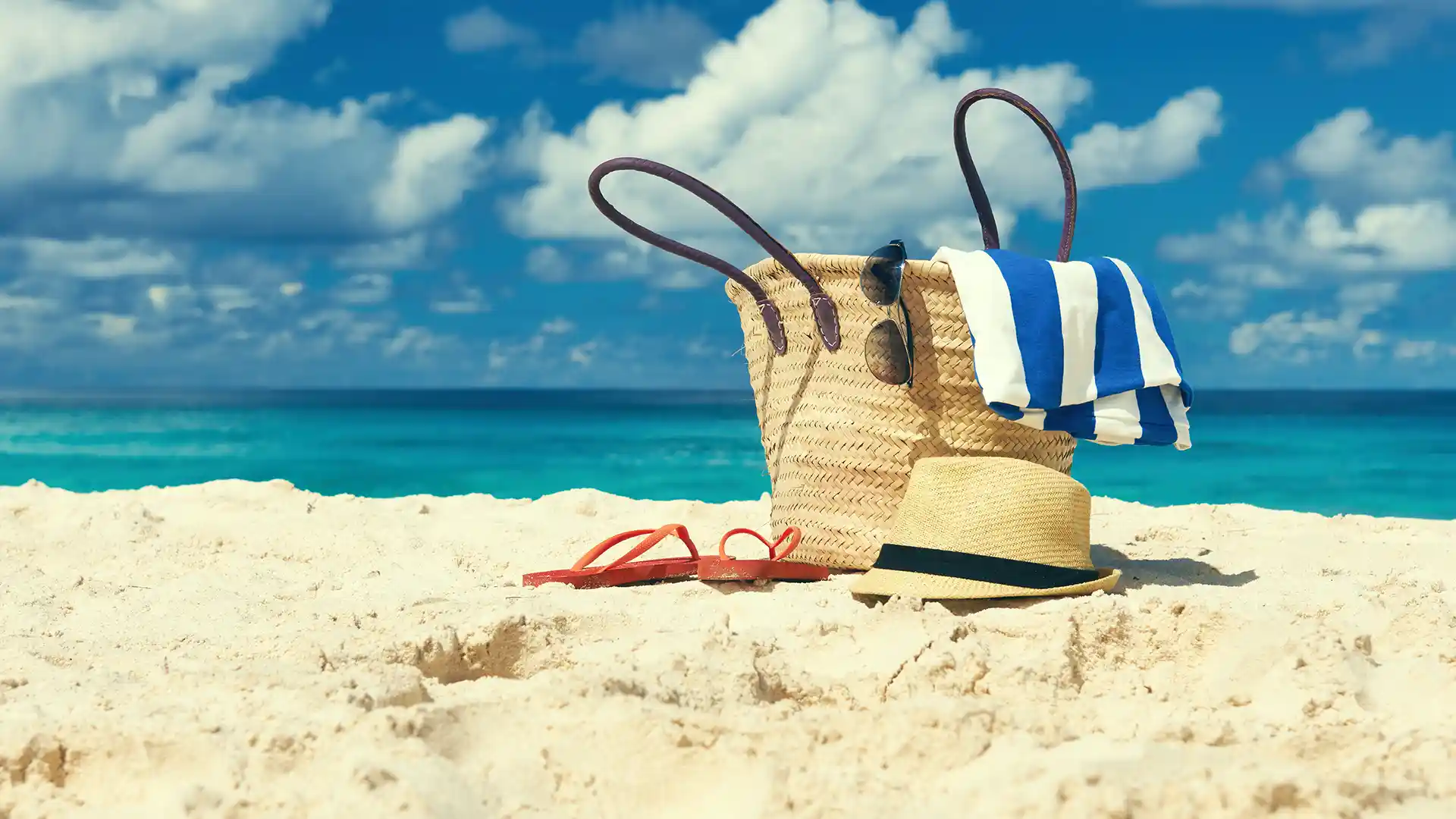 Beige carry-on bag in the sand with a hat and flip flops next to it and blue ocean water in the background.