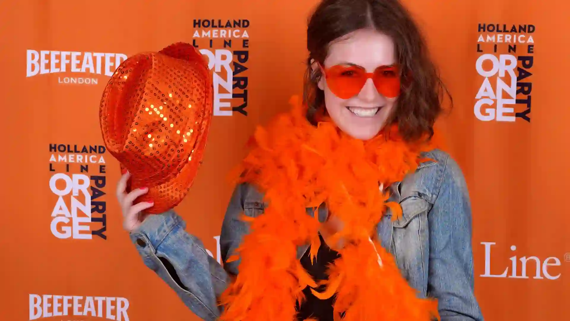View of person wearing orange sunglasses and boa with hat, standing in front of Orange Party background.