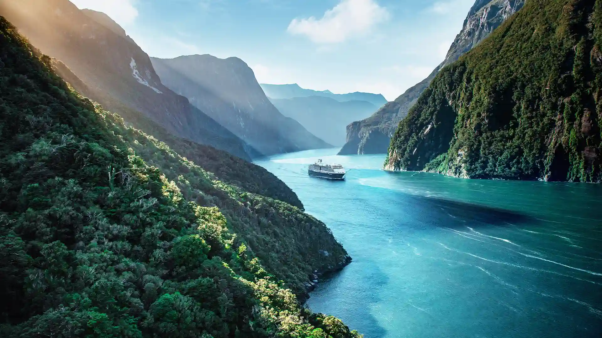 View of Holland America Line cruise ship sailing across the Milford Sound in New Zealand, surrounded by lush green landscape.