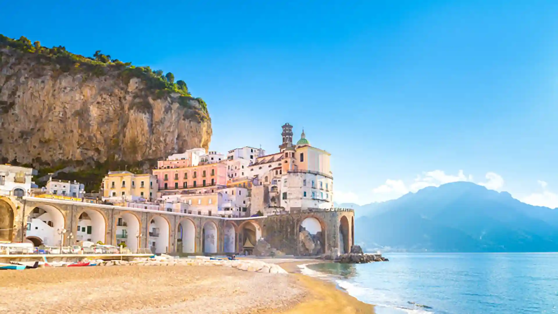 View of a sandy shore along the Amalfi Coast in Italy with buildings and lush landscapes in the background.