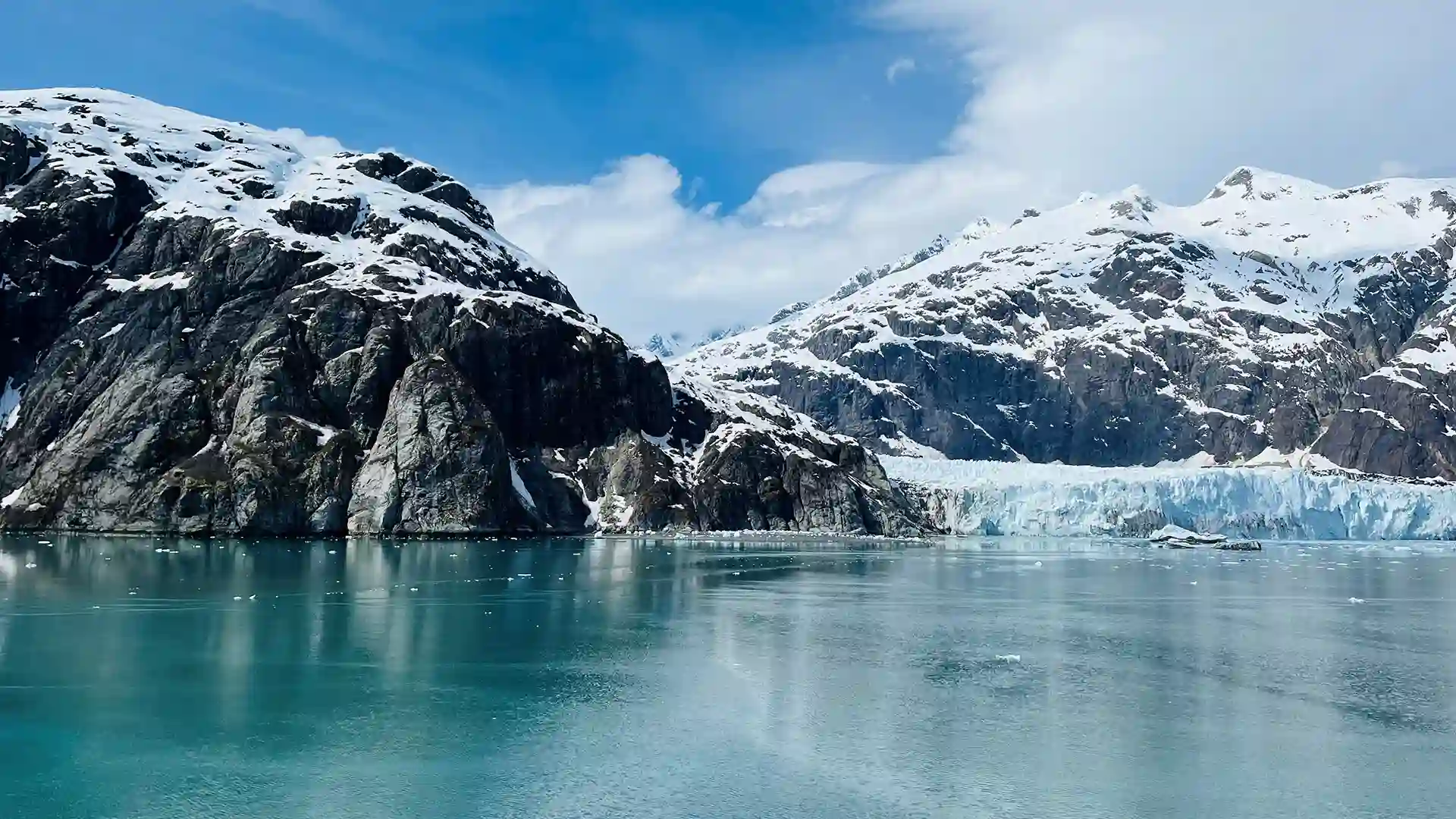 View of snowcapped mountains and glacier in Glacier Bay, Alaska, beneath partly cloudy skies.