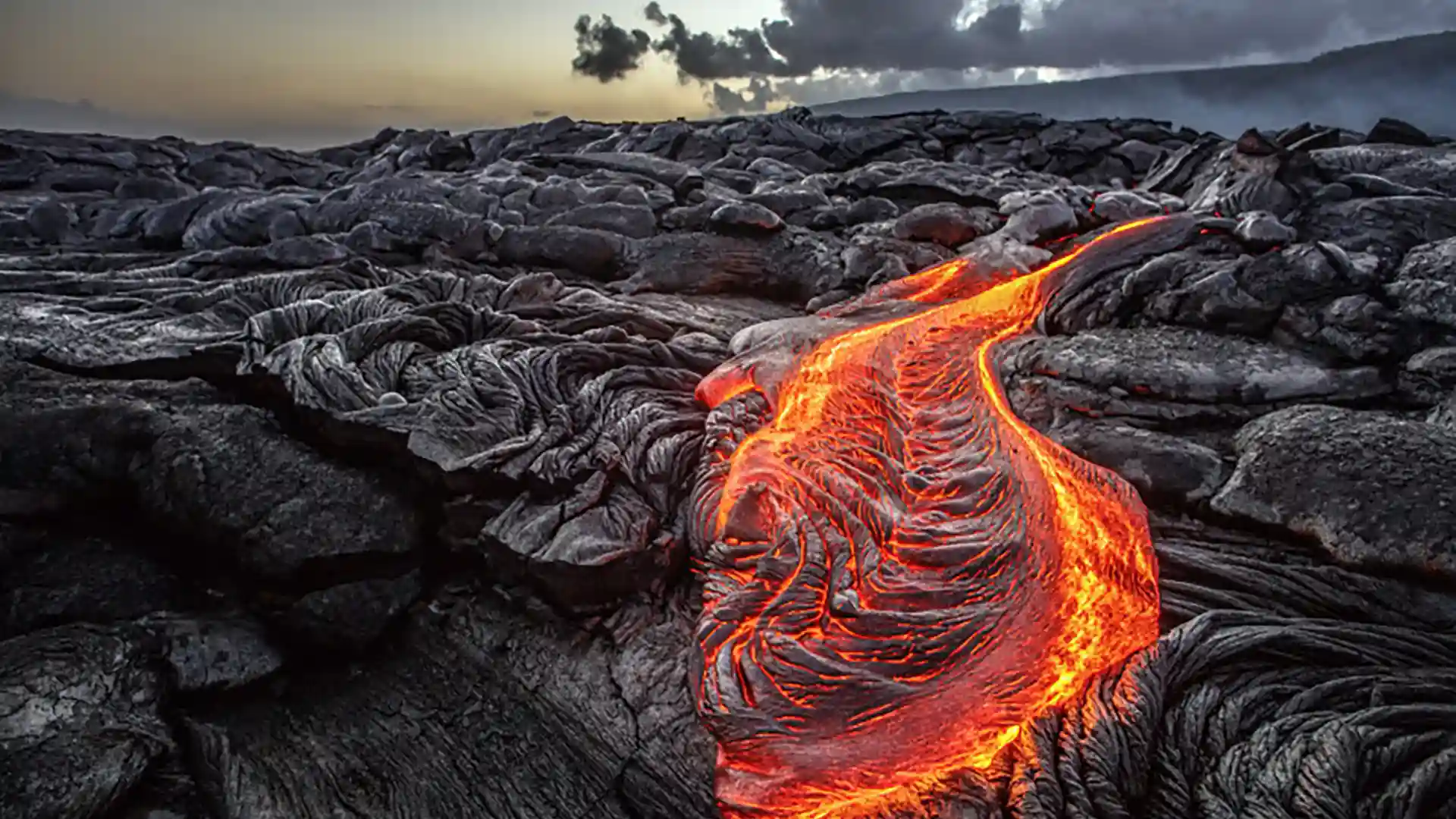 View of orange lava flowing across black rocky terrain on volcano in Hawaii.