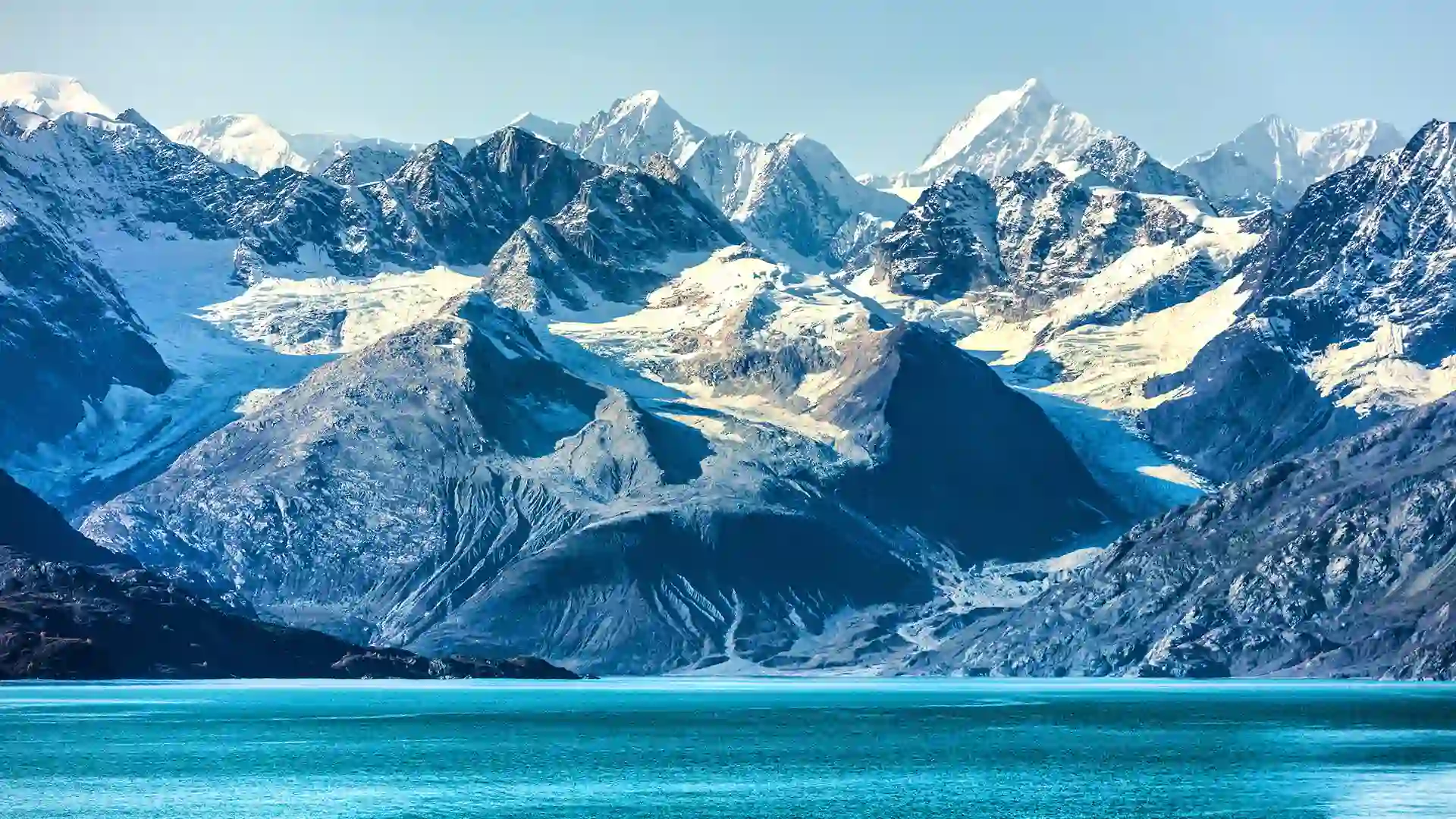 View of Glacier Bay National Park in Alaska, with its snowcapped towering mountain range, along the bright blue shoreline.