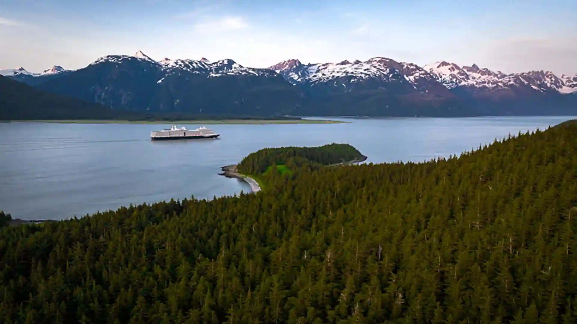 View of Holland America Line cruise ship sailing around Alaska near lush forests and snowcapped mountains.