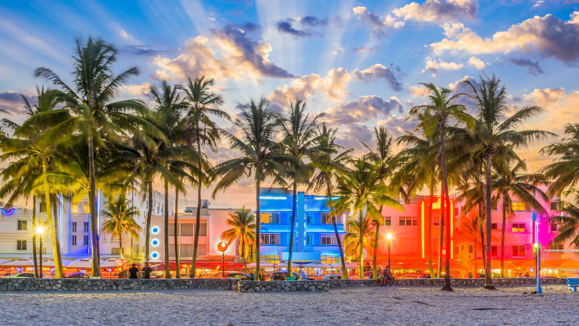 Palm trees and buildings line up along Miami’s dynamic waterfront with blue, pink, and orange lights.