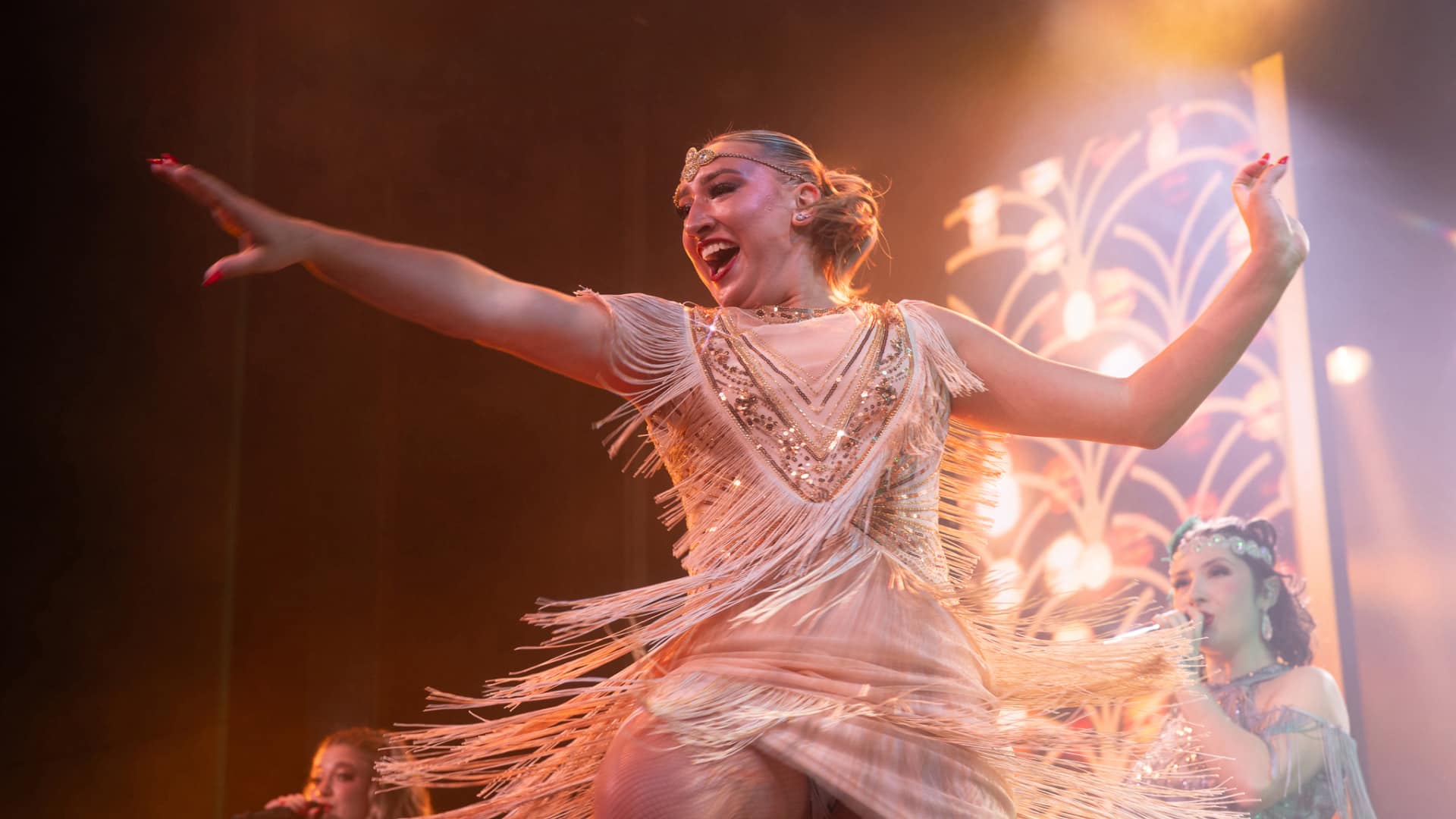 Performer in a fringed costume on stage with decorative lighting and musicians in the background.