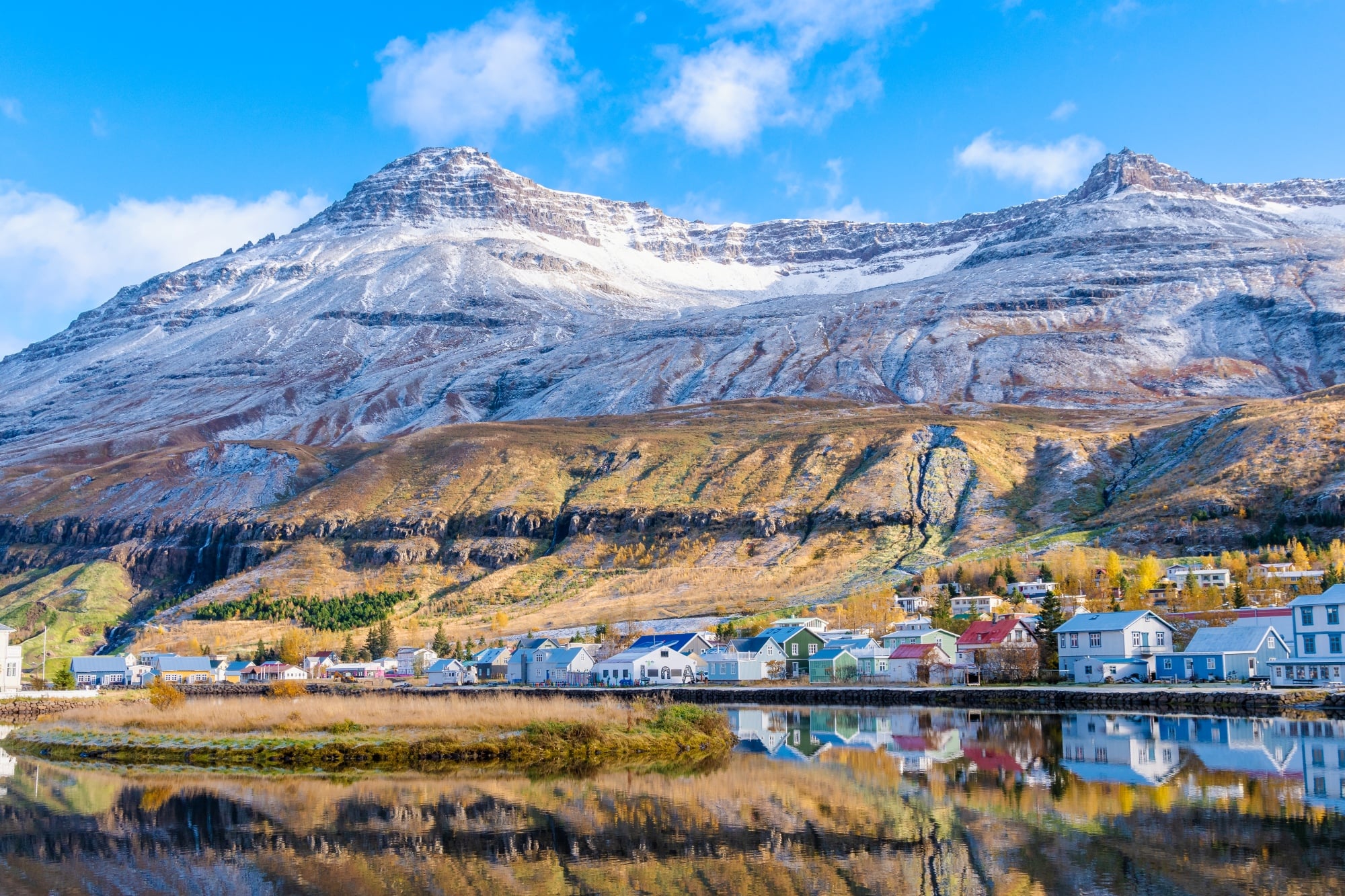 Colorful houses lined up on clear water with a snowy mountain in the background.