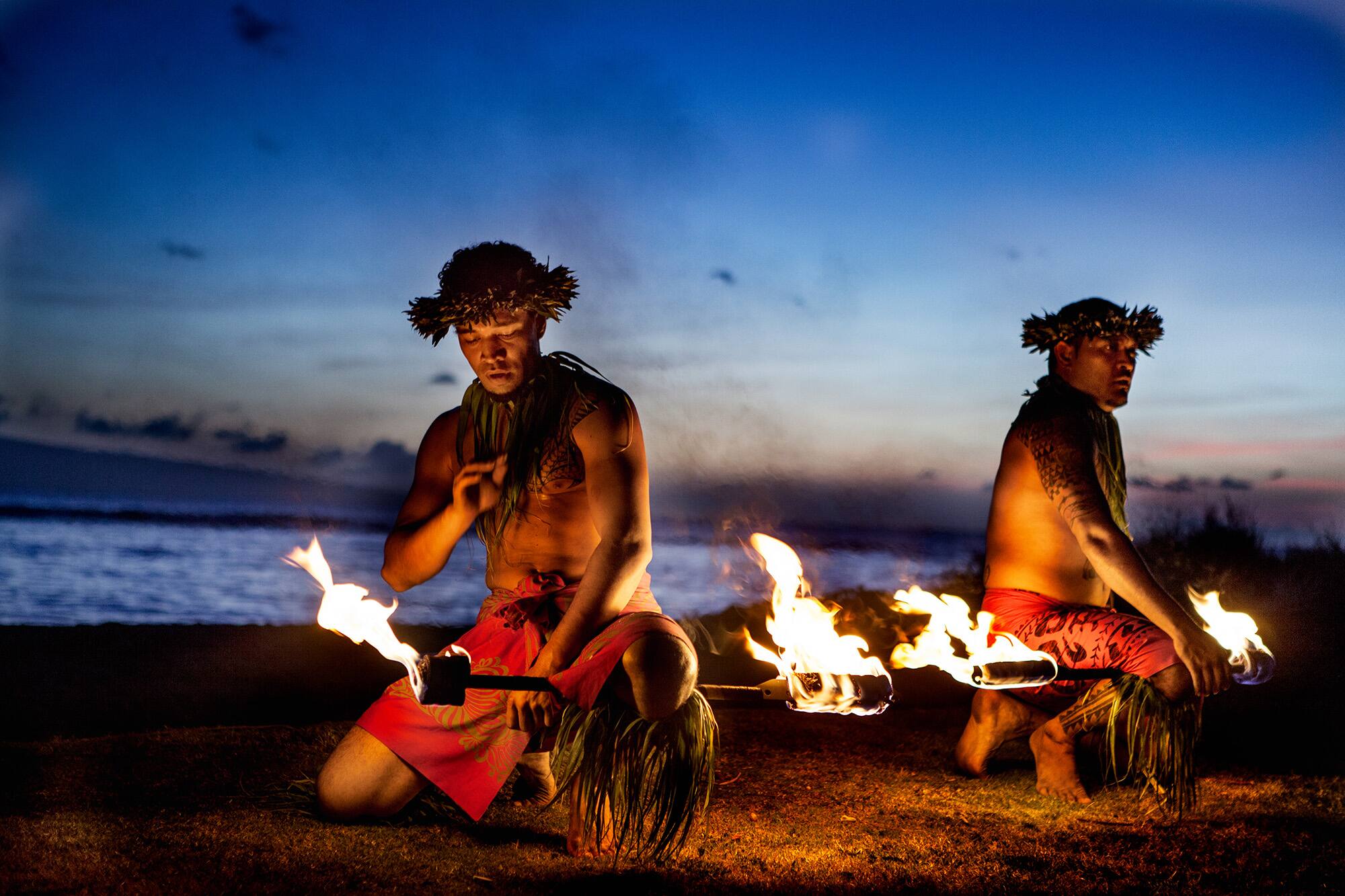 Two male Hawaiian fire dancers holding lit fire sticks on a beach.