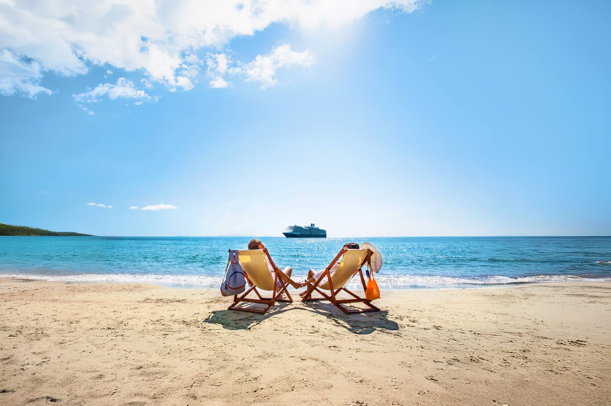 A couple in beach chairs holding hands on a Caribbean island with a Holland America cruise ship on turquoise water in the background.
