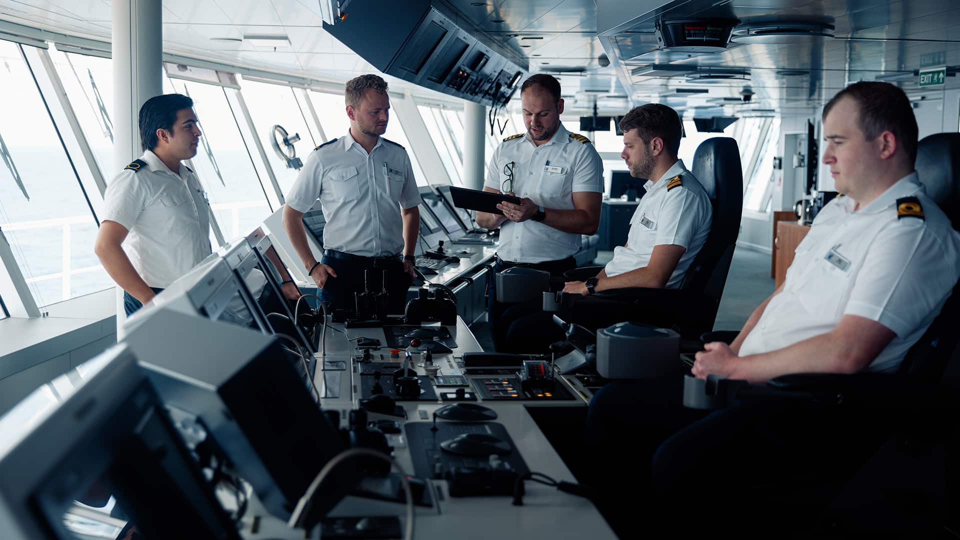 Five cruise ship officers wearing white shirts and navy blue pants at work in the Bridge.