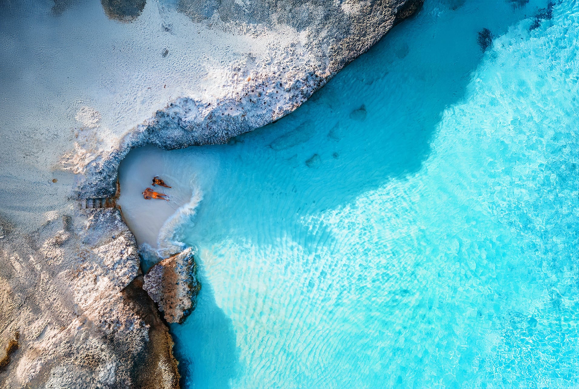 A man and woman in orange swimwear from above on a small private beach in front of the ocean.