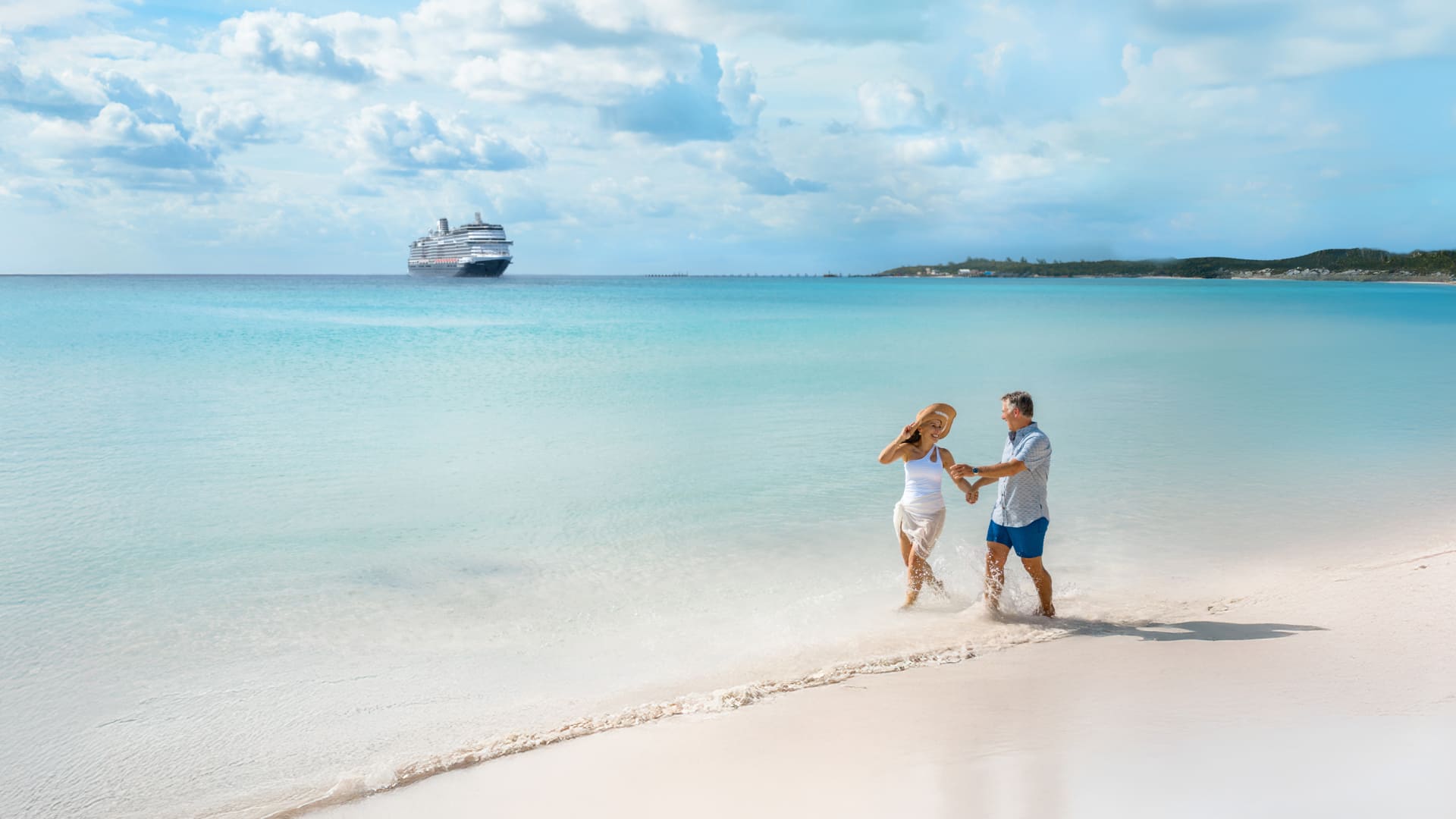 A man and woman frolic in the Caribbean’s white sand with a Holland America ship in the background.