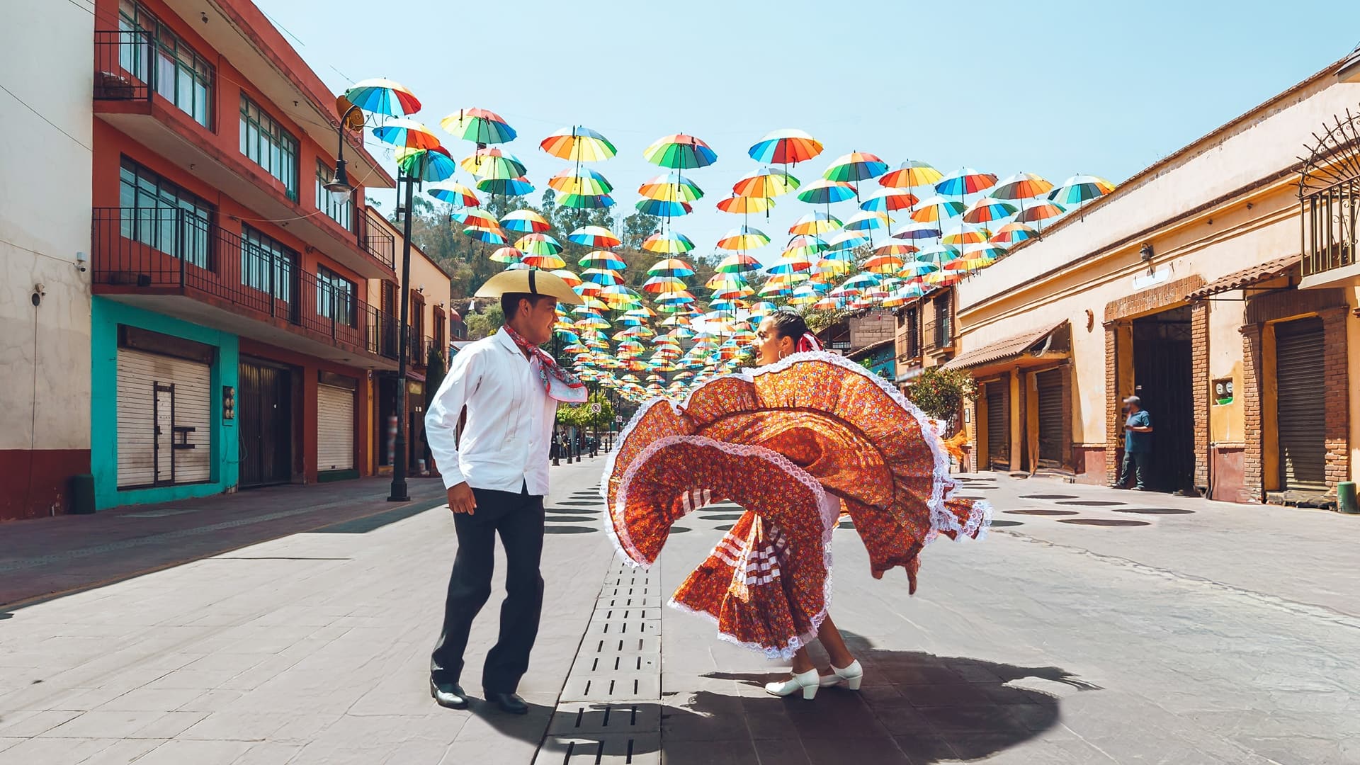 A man in a cowboy hat and woman in a flowing orange skirt and white heels dancing on a street beneath colorful umbrellas.
