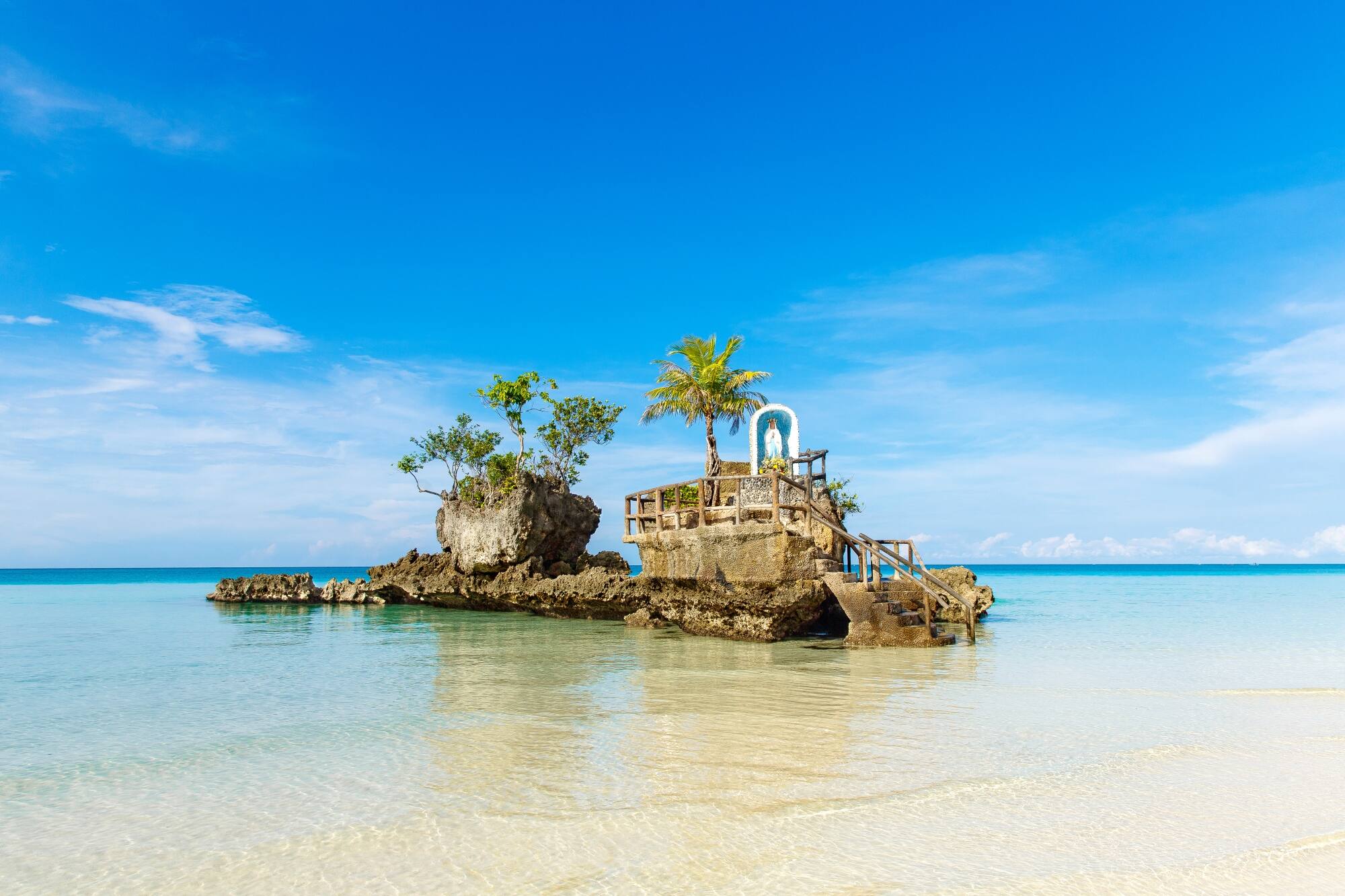 Rock formation with a small shrine and plants surrounded by clear shallow water.