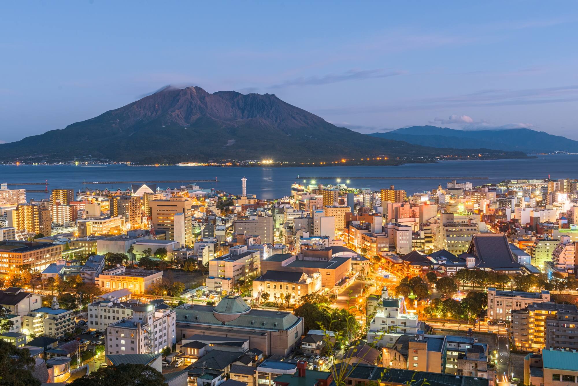 City skyline along the waterfront with a large volcano in the background at dusk.
