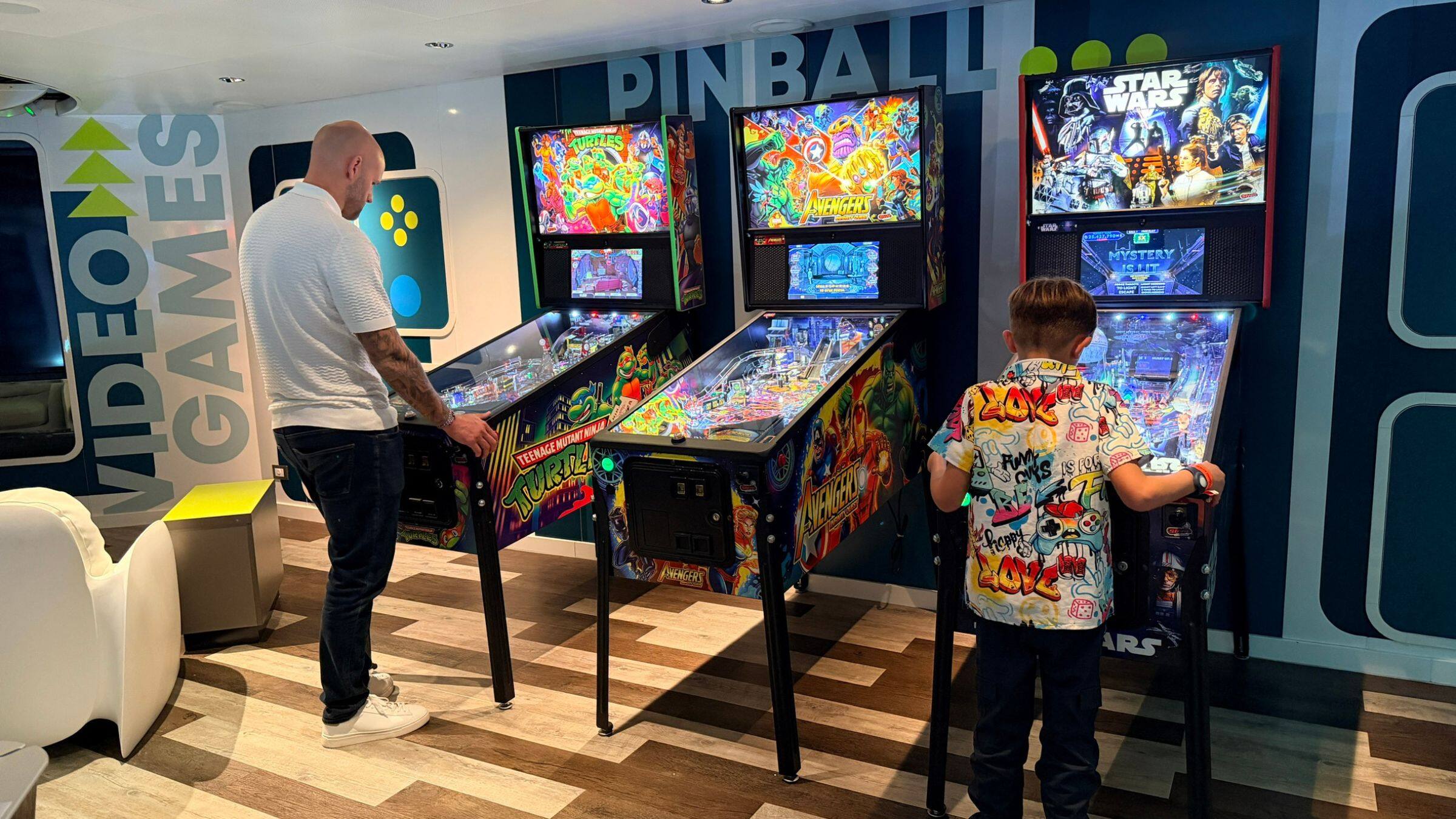 A man in a white t-shirt and a boy in a colorful shirt play pinball in Hang10, cruise ship arcade.
