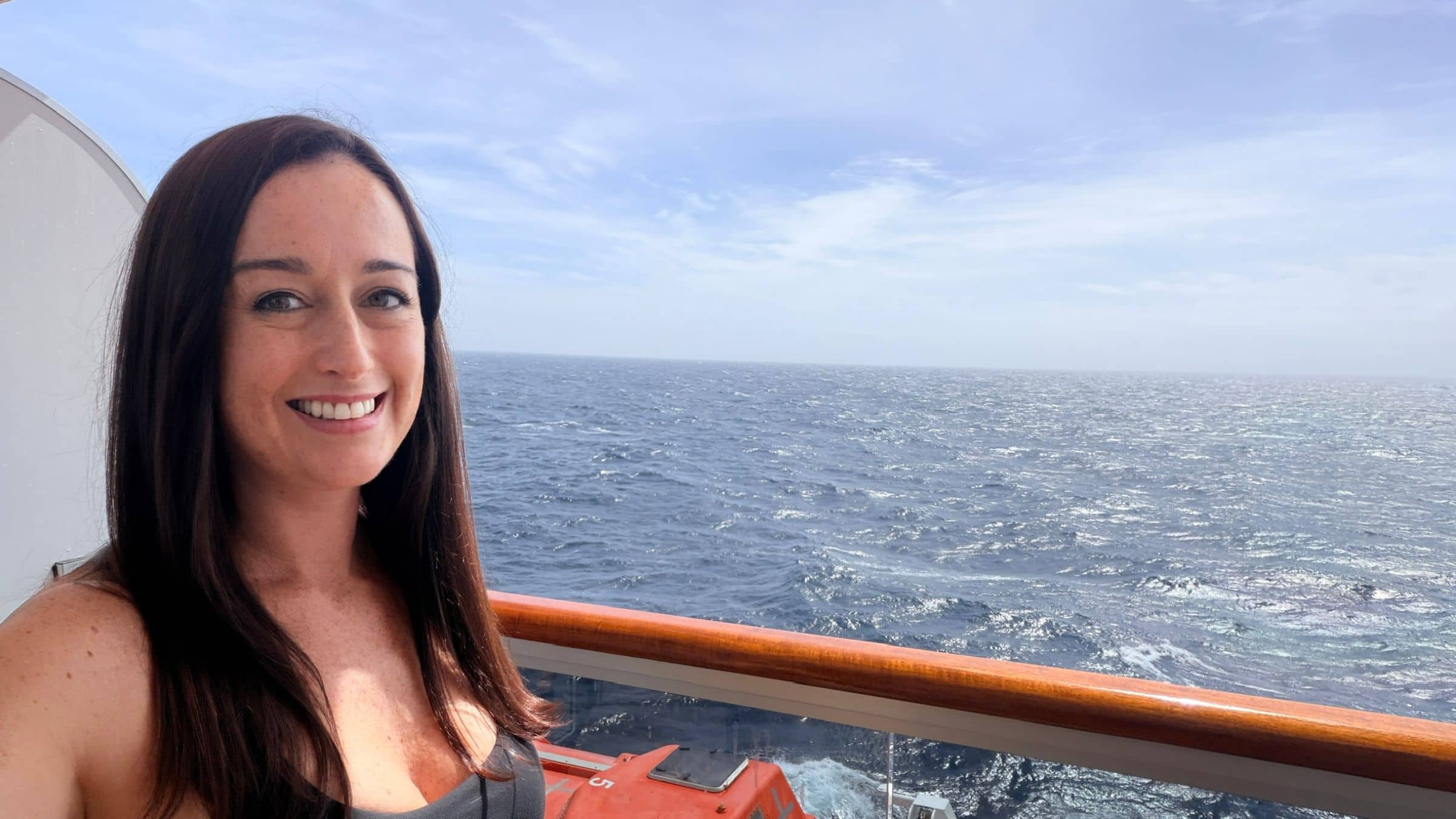 A woman with shoulder length brown hair wearing a tank top on the deck of Nieuw Statendam wiith ocean views behind her.
