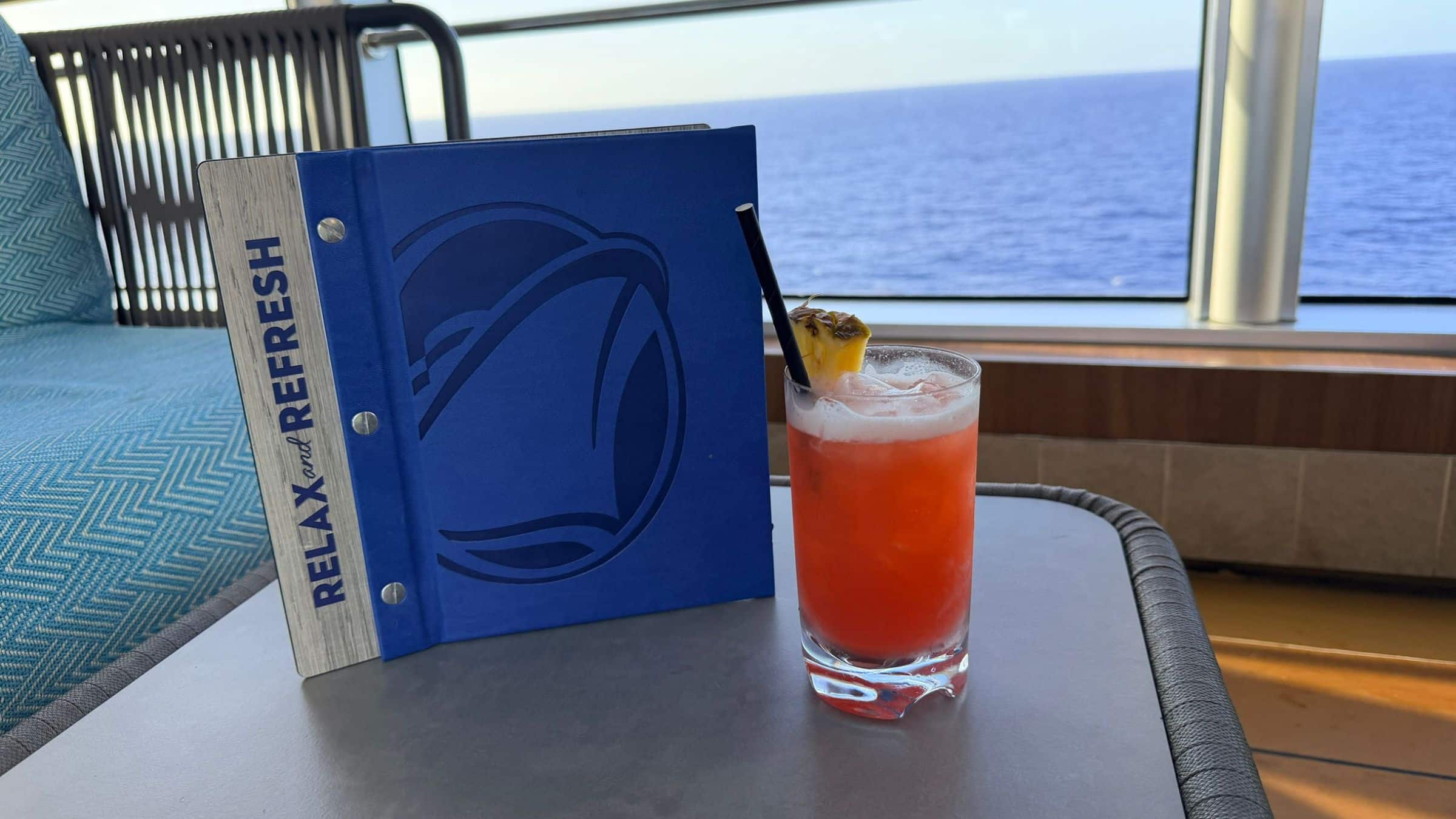 A blue Holland America menu positioned upright with the text Relax & Refresh next to a orange drink with a pineapple garnish in front of a window.