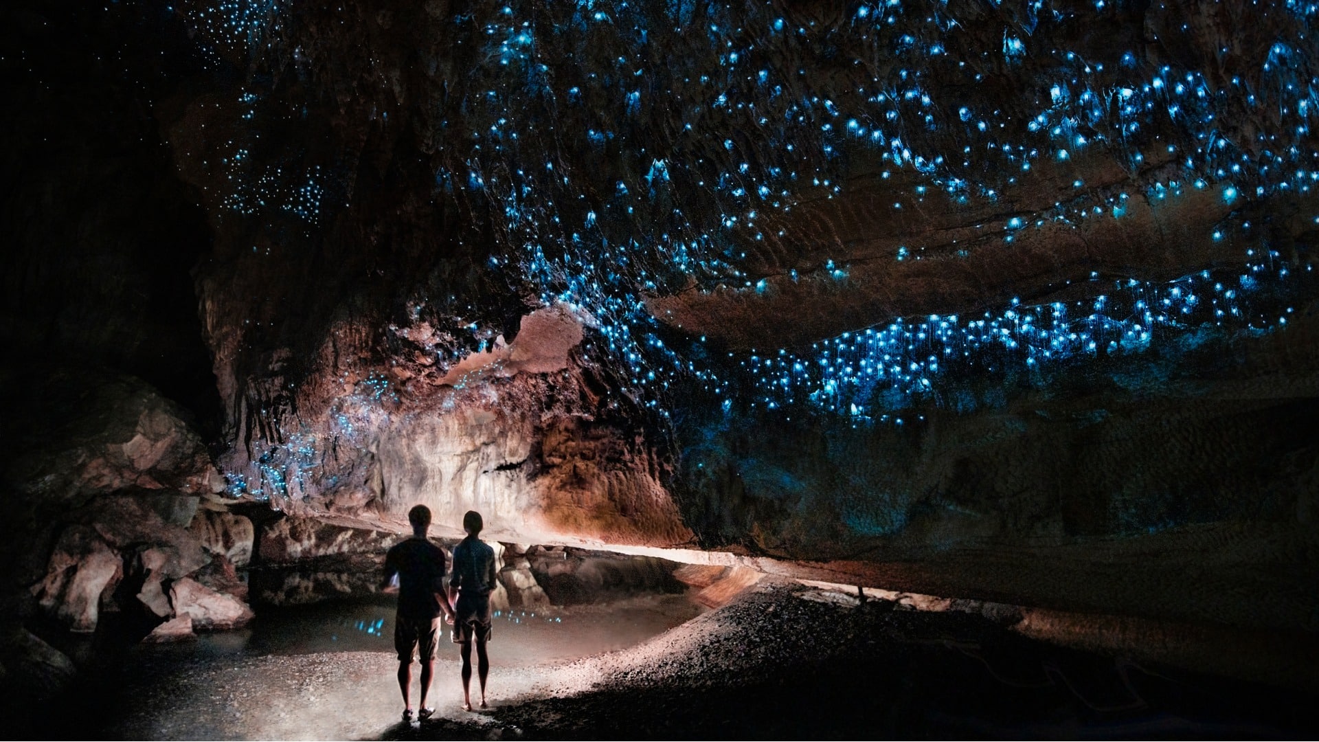 A man and women stand in New Zealand’s Waitomo Cave under the blue lights of glowworms.
