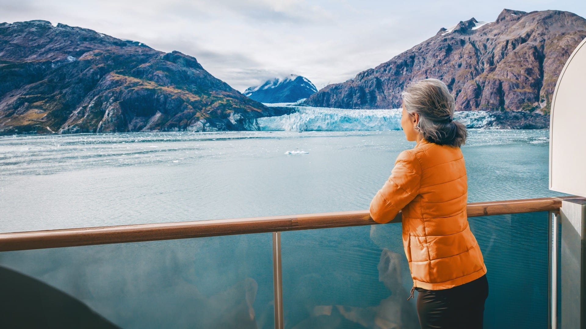 Person standing on a cruise ship balcony looking at a glacier in a mountain landscape.